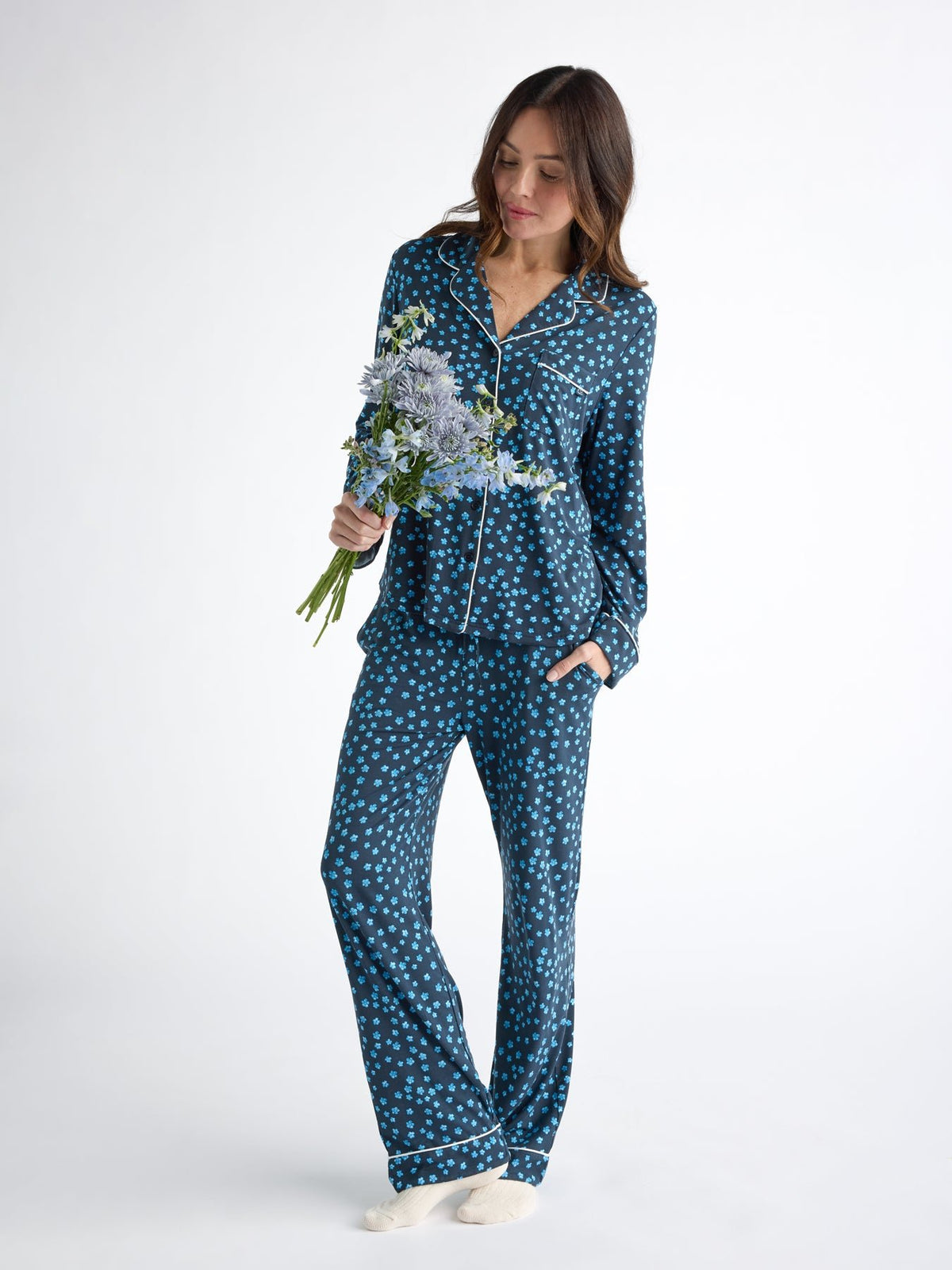 A person in Cozy Earth Women's Bamboo Stretch-Knit Pajama Pant with blue polka dots and white piping stands barefoot against a white background, holding a bouquet of flowers. 