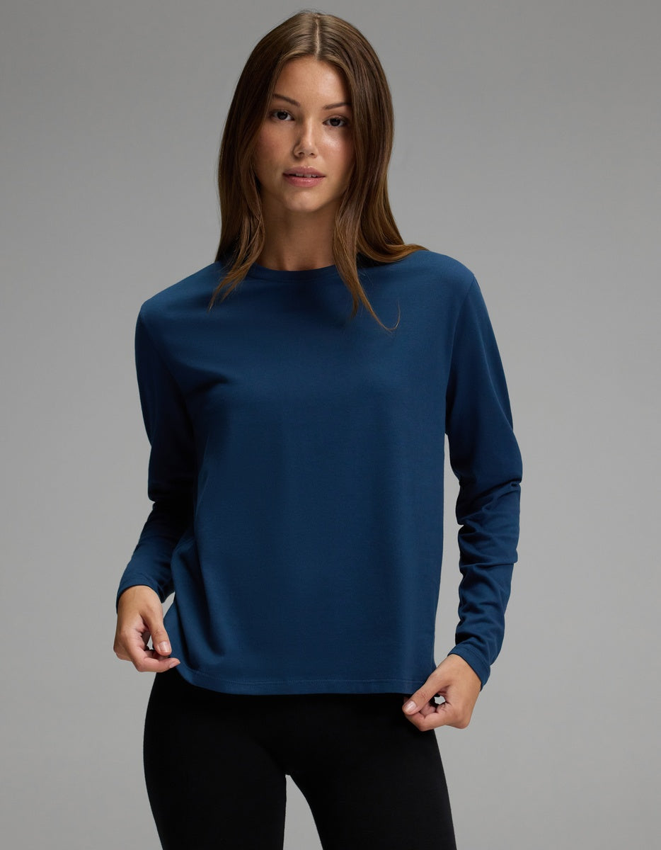 A person with long brown hair is wearing the Cozy Earth Women’s All Day Long Sleeve Tee in navy blue and black pants, standing against a plain gray background and looking at the camera.
