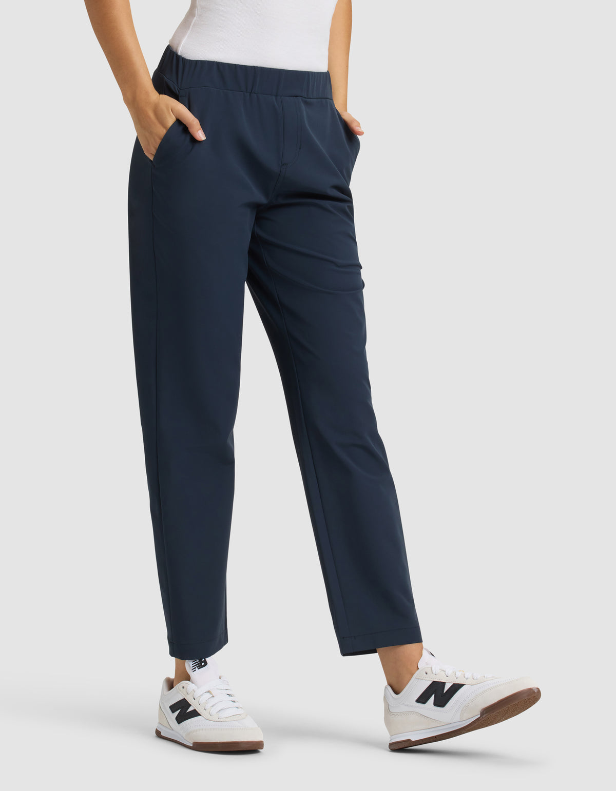 A person wears Cozy Earth Women's Always Cropped Pant in navy blue with a white top and white sneakers with brown soles. Hands are in the pants' pockets, standing against a plain light gray background. 