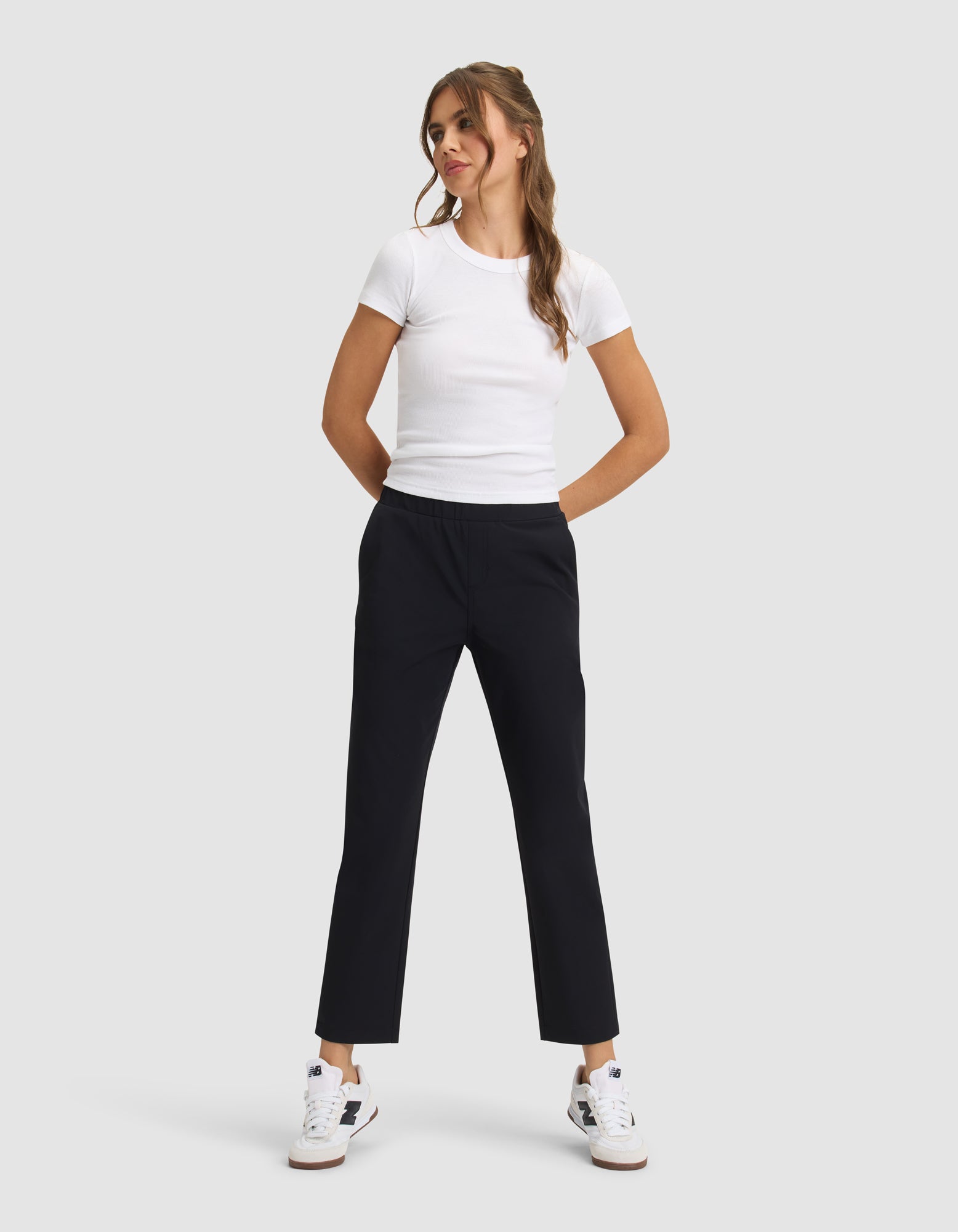 A woman stands against a light gray background, smiling with her hands in her pockets, wearing Cozy Earth's Women's Always Cropped Pant in black, a fitted white t-shirt, and white sneakers with blue and tan accents. |Color:Jet Black