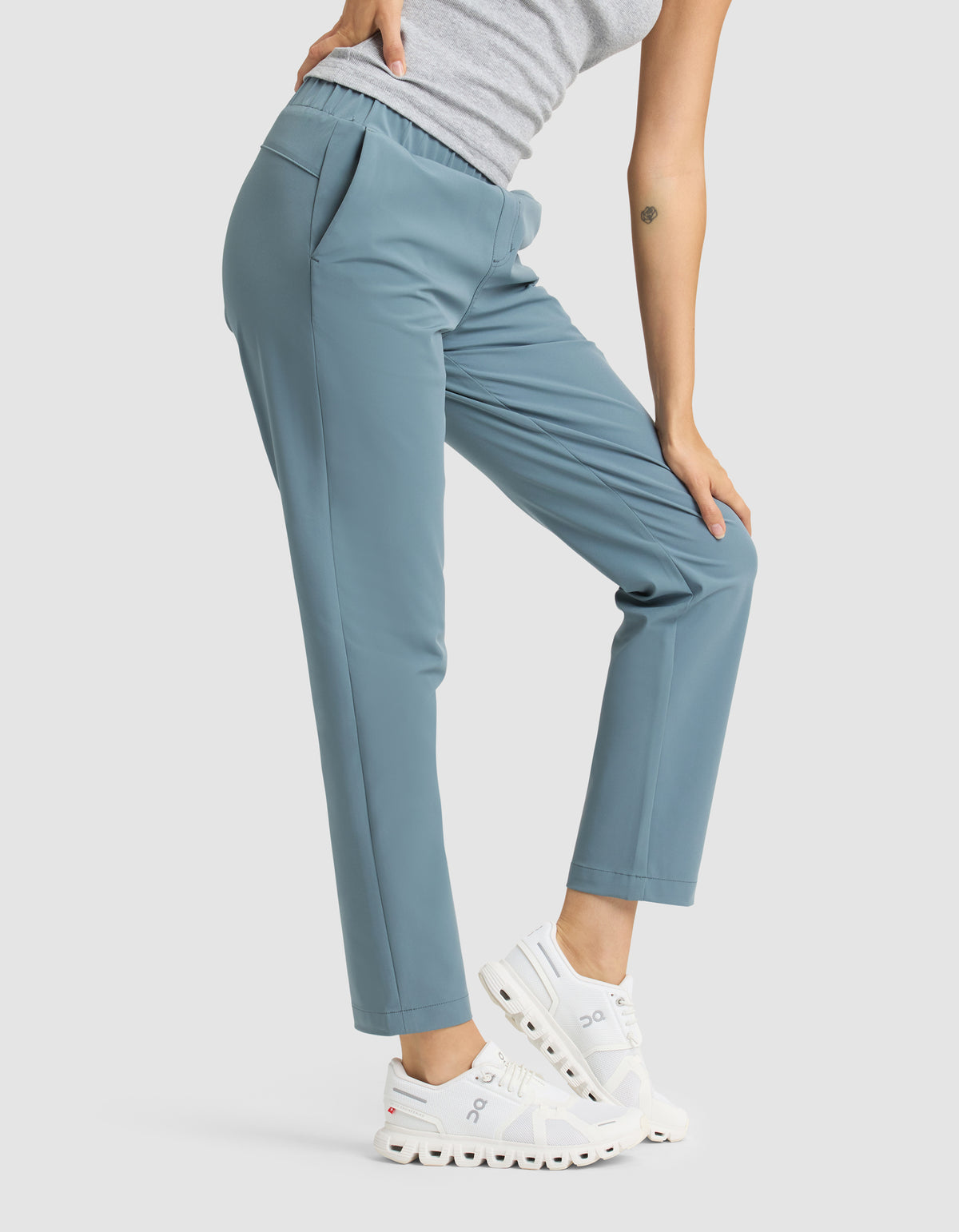 A person wearing Cozy Earth's Women's Always Cropped Pant in blue, paired with a grey sleeveless top and white sneakers, stands with one knee bent and a hand resting on their thigh. 