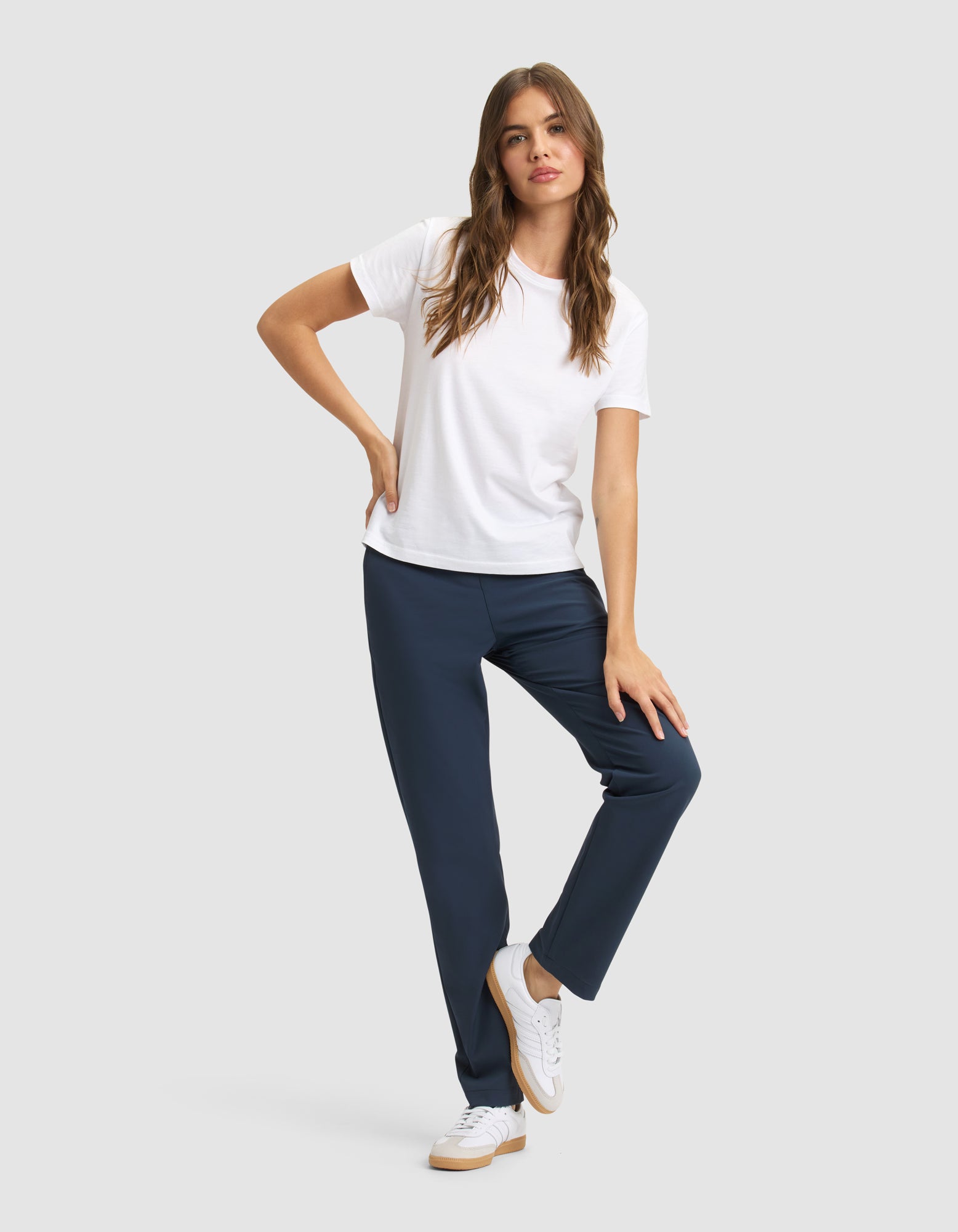 A woman with long, wavy brown hair poses against a plain background, wearing Cozy Earth's Women's Always Pant in navy blue, a white T-shirt, and white sneakers, with one hand on her hip and one leg slightly bent. |Color:Eclipse