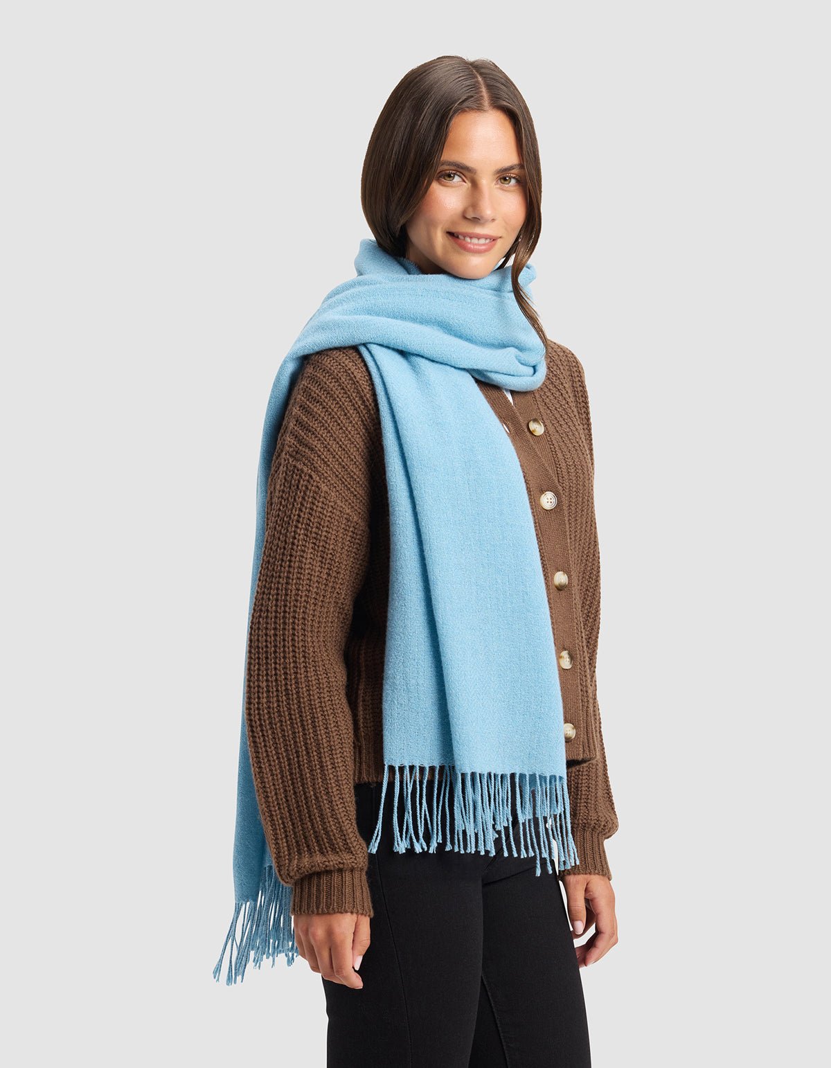 A woman with shoulder-length brown hair smiles while standing against a plain light gray background, wearing Cozy Earth's Amelia Wrap in brown over black pants and a light blue fringed scarf draped around her neck.