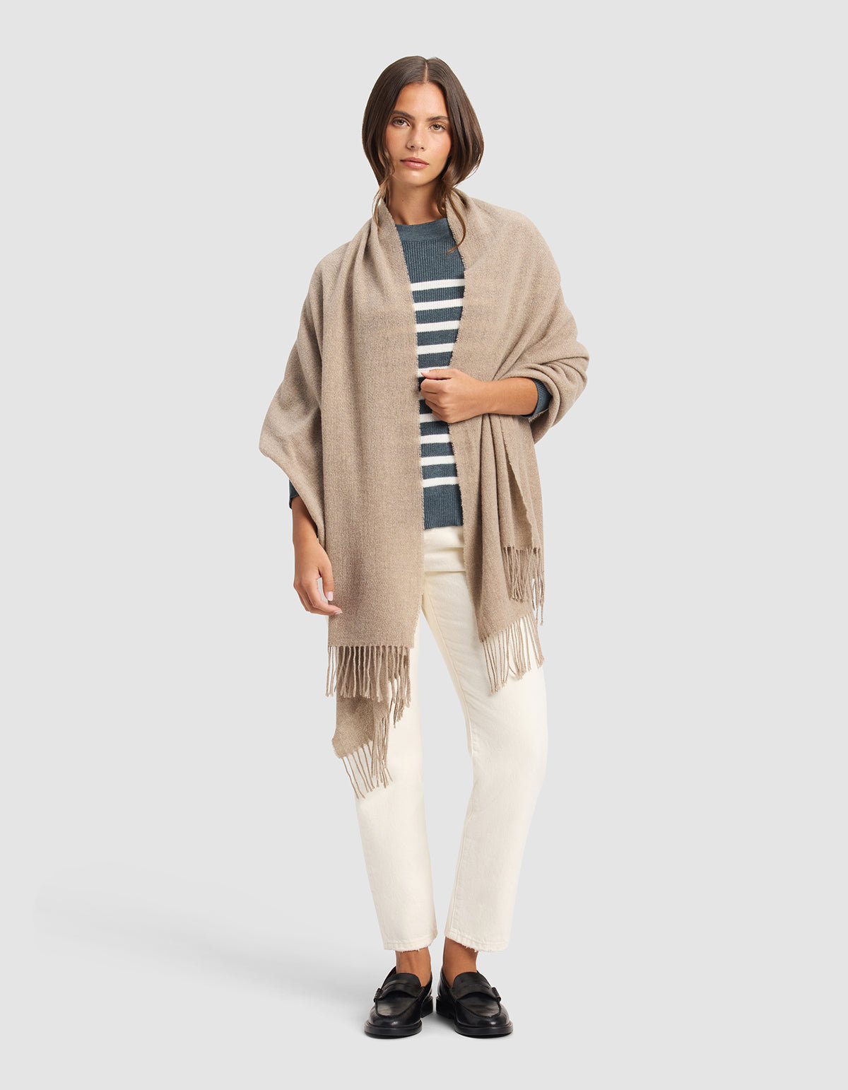 A woman with straight brown hair and a neutral expression stands against a plain background wearing the Cozy Earth Amelia Wrap, a navy and white striped sweater, off-white pants, and black loafers.