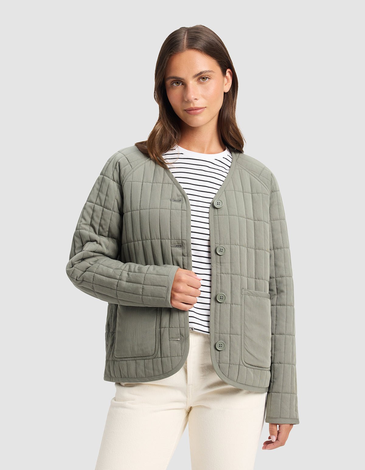 A woman wears the Cozy Earth Women's Andie Quilted Jacket in light green over a white and black striped shirt with cream-colored pants, standing against a plain light gray background.