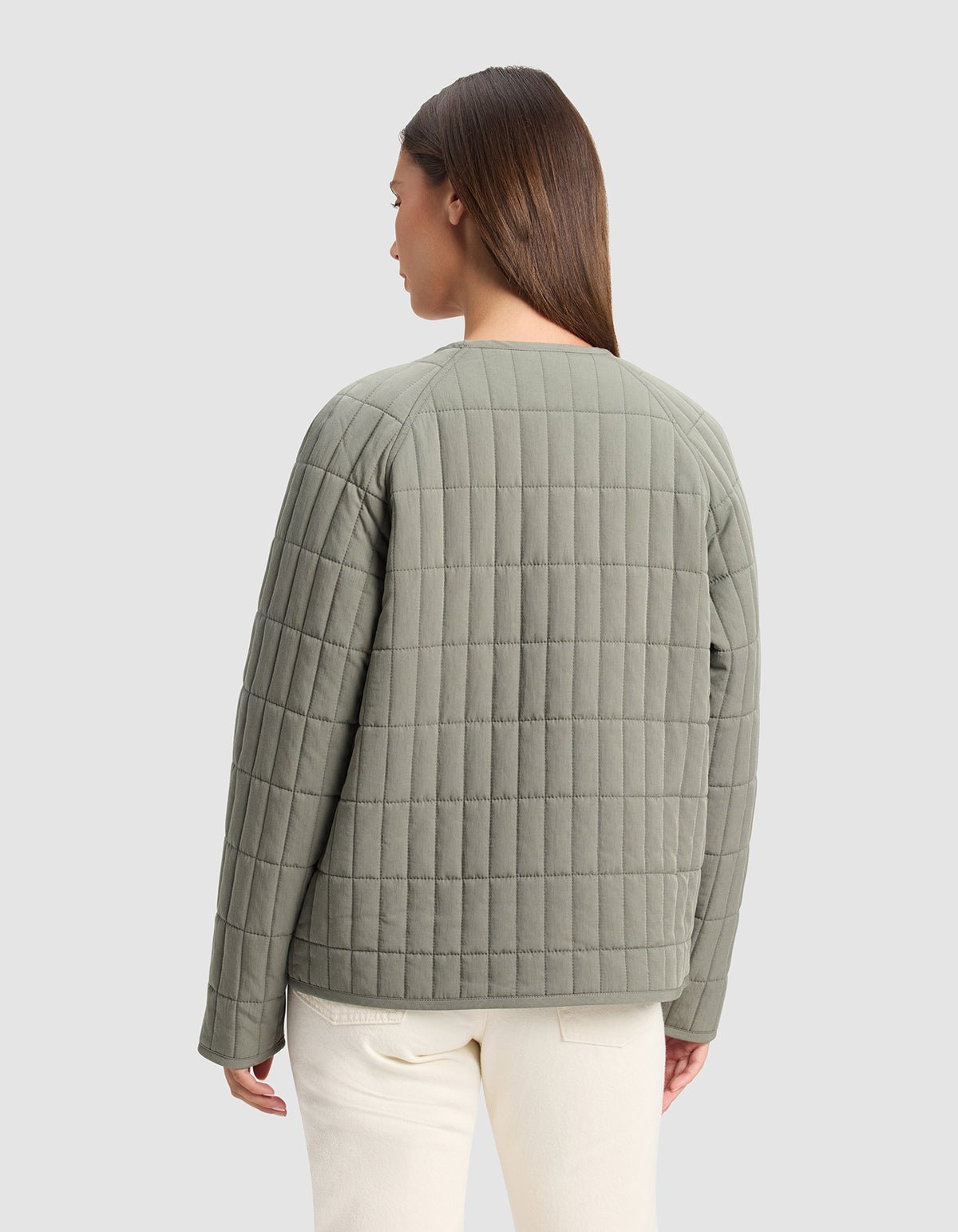 A woman with straight brown hair, facing away from the camera, wears the Cozy Earth Women's Andie Quilted Jacket in light olive green and off-white pants against a plain light gray background.