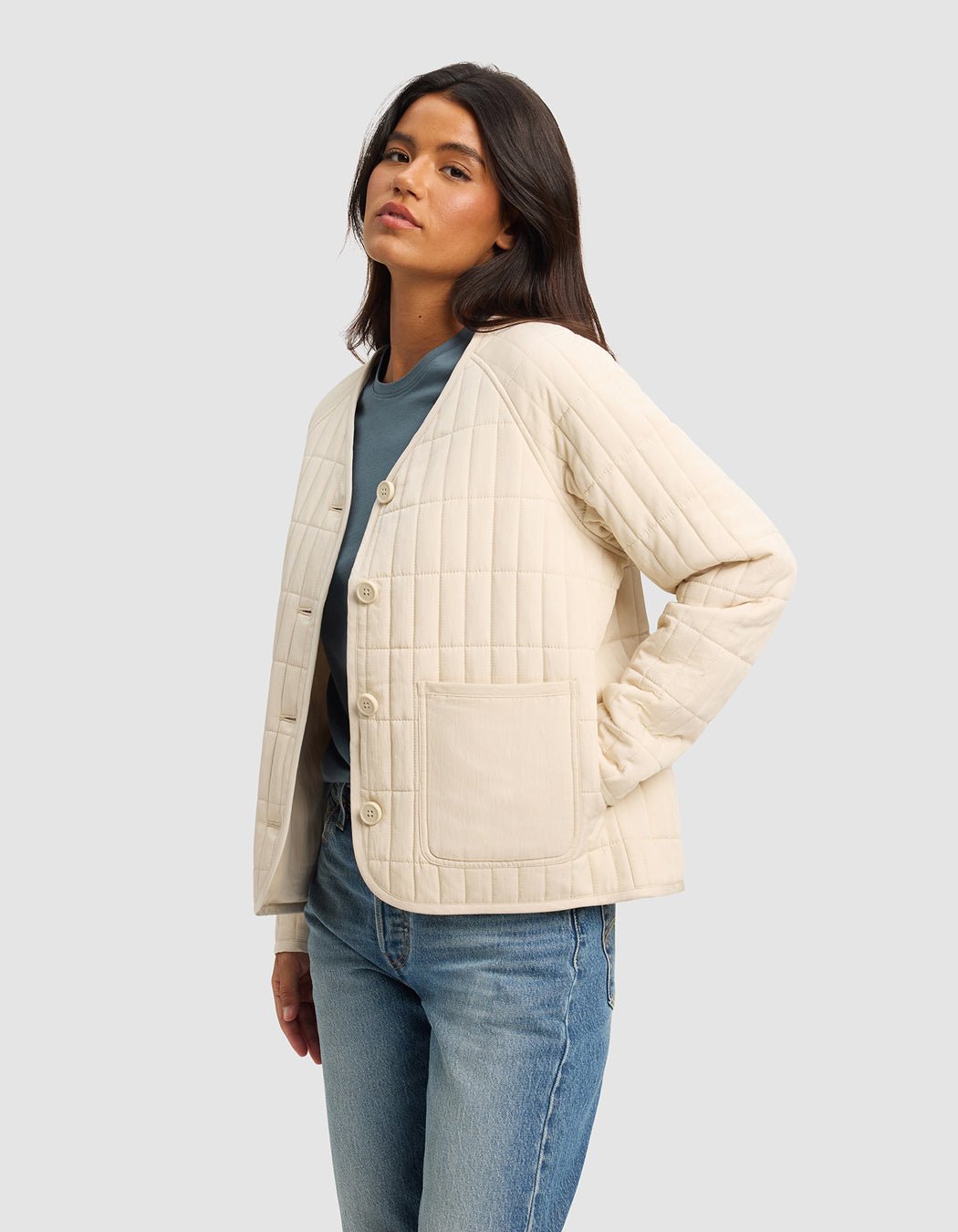 A woman with long dark hair wears the Cozy Earth Women's Andie Quilted Jacket in cream over a blue top and light blue jeans, standing with one hand in her pocket against a plain light gray background.