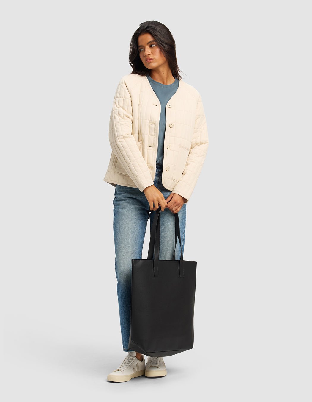 A woman with dark hair wears the Cozy Earth Women's Andie Quilted Jacket in cream over a blue shirt and jeans, paired with white sneakers. She stands against a light gray background, holding a large black tote bag.