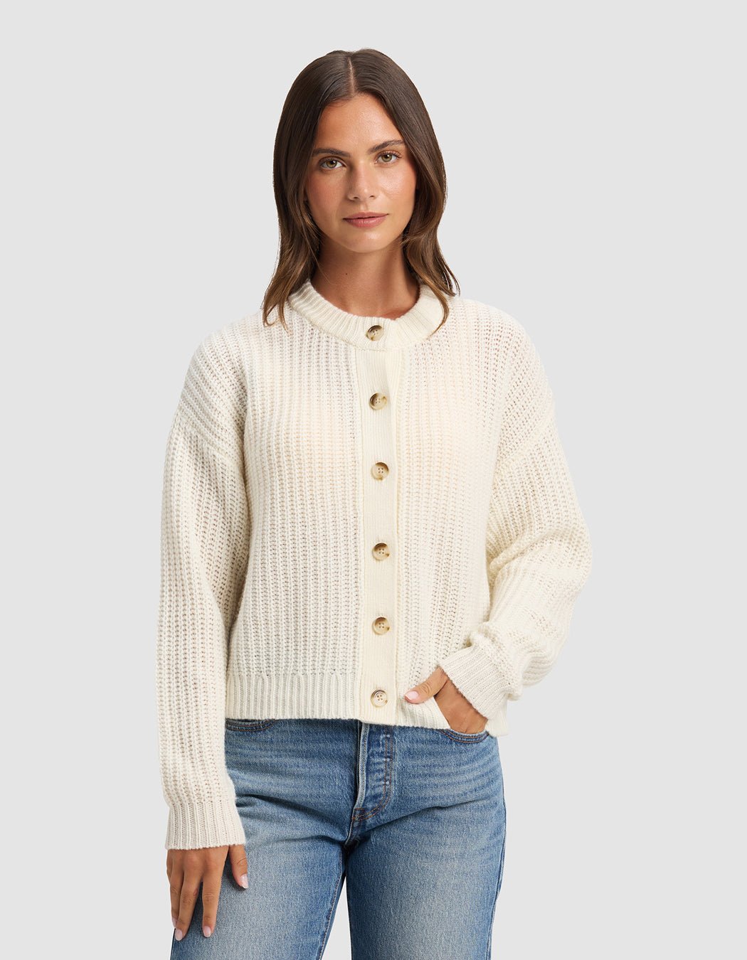 A woman with straight brown hair wears the Cozy Earth Women's Arden Cardigan in cream and blue jeans, standing against a plain light gray background, facing forward with one hand in her pocket.|Color:Bone