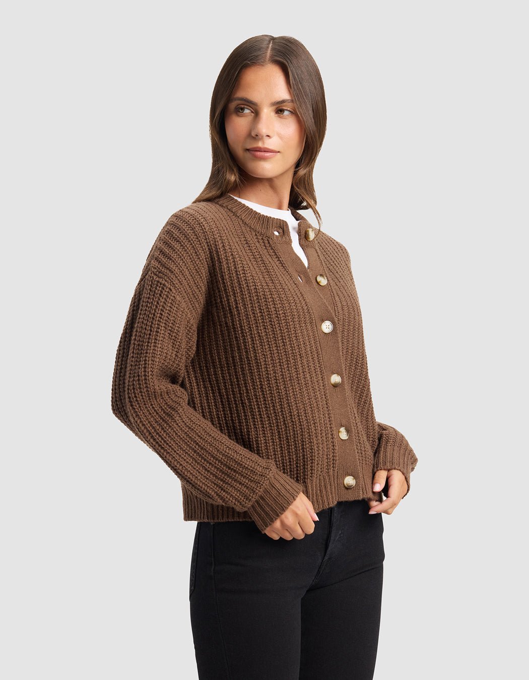 A woman with long brown hair models the Cozy Earth Women's Arden Cardigan, a brown ribbed button-up, layered over a white top and black pants against a plain light gray background.