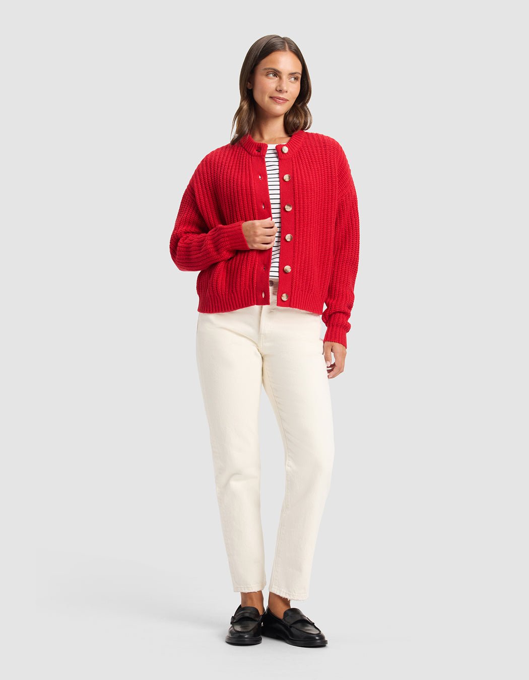 A woman with straight brown hair poses against a plain background in a Cozy Earth Women's Arden Cardigan (red), layered over a striped shirt, paired with cream pants and black loafers. She displays a relaxed, neutral expression.