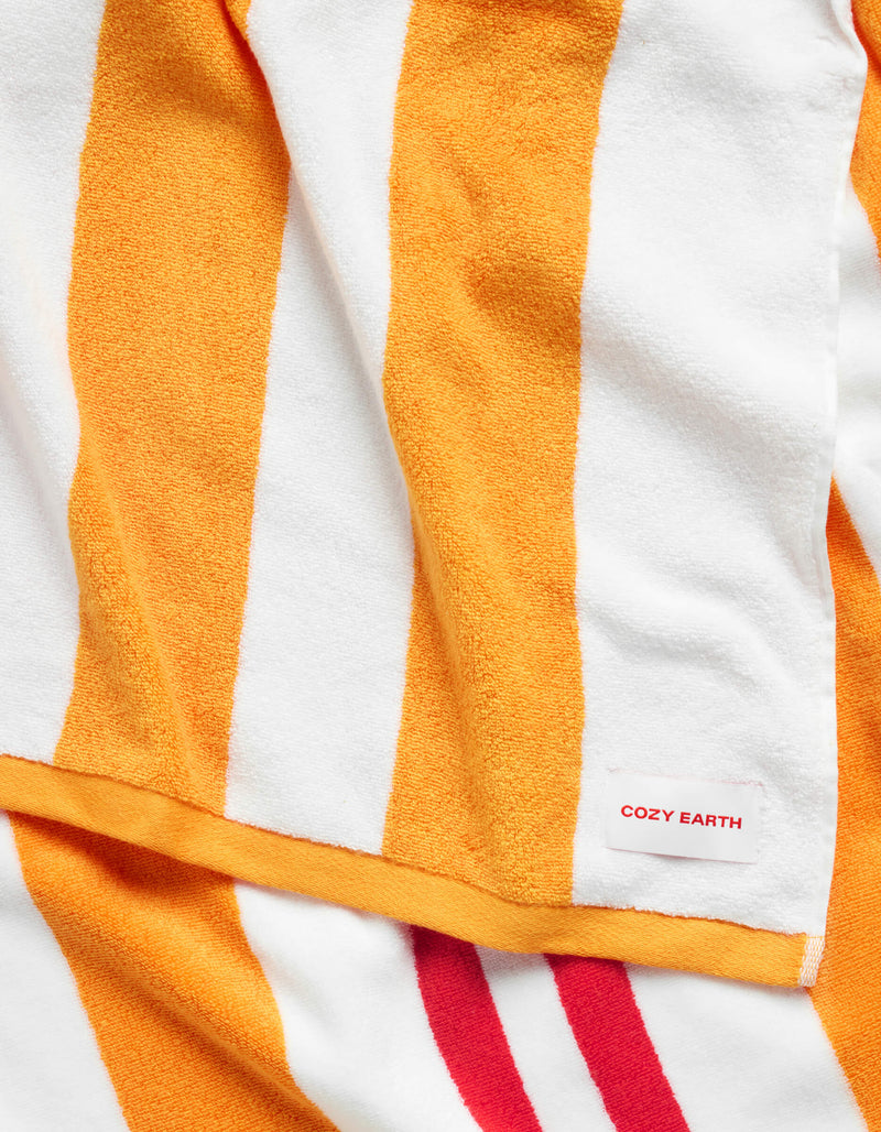 A close-up of the Classic Stripe Resort Towel by Cozy Earth shows its wide orange and white stripes, with a small white "Cozy Earth" tag visible in the corner.