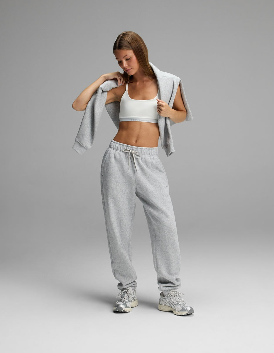 A person wearing a white Cozy Earth Women’s Balance Bra, gray sweatpants, and a gray sweatshirt draped over their shoulders.