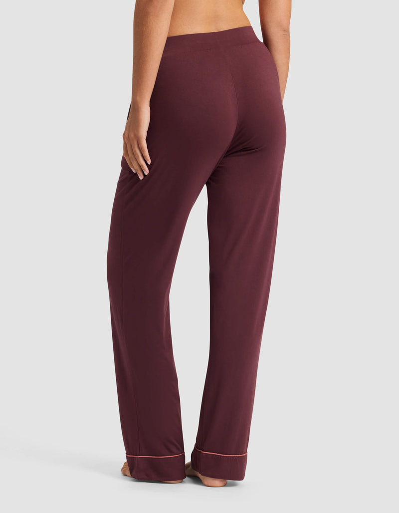 A person stands barefoot on a white background, wearing Cozy Earth's Women's Bamboo Stretch-Knit Pajama Pant in burgundy with a relaxed fit and light-trimmed cuffs; their back and legs are visible from mid-back to feet.
