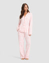 A woman poses barefoot against a light gray background wearing Cozy Earth's Women's Bamboo Stretch-Knit Long Sleeve Pajama Set in a light pink and white pattern, featuring a button-up top and matching pants. |Color:Tulip Toile