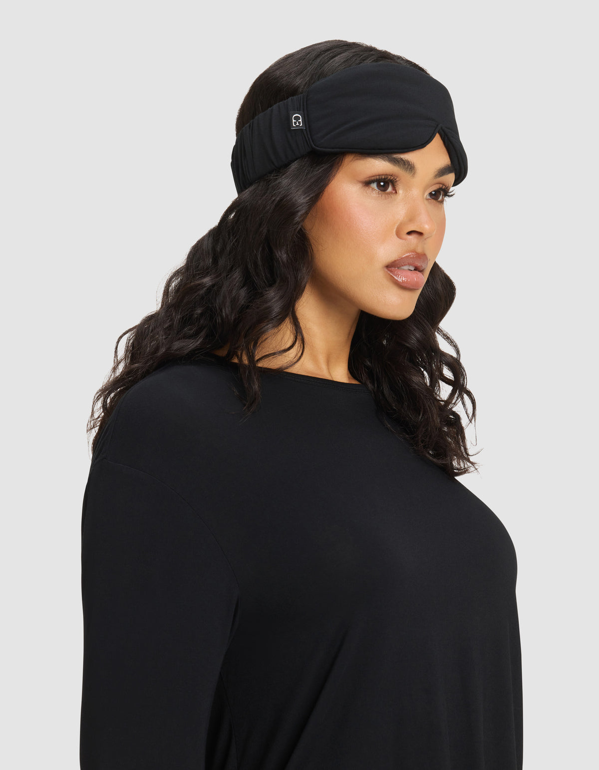 Woman with long wavy dark hair wears the Cozy Earth Bamboo Stretch-Knit Sleep Mask, paired with a loose black top, gazing slightly to the side against a plain light gray background.