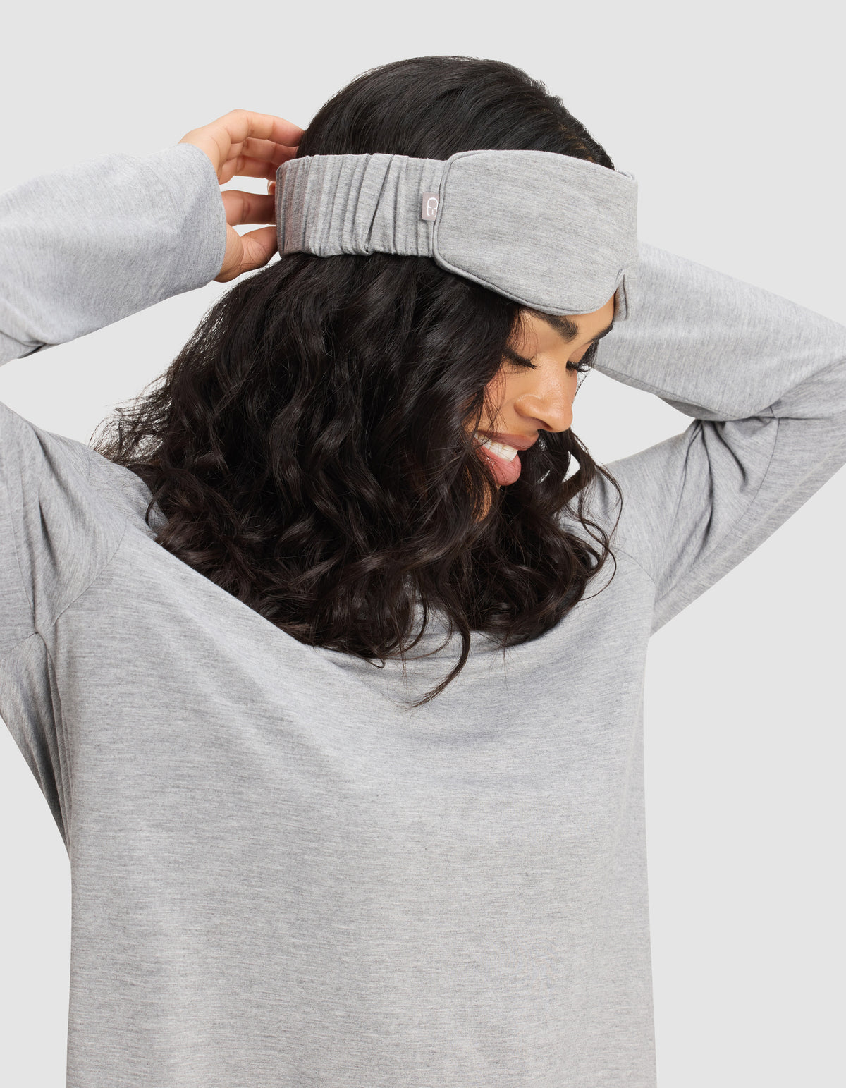 A woman with wavy dark hair smiles while adjusting the Cozy Earth Bamboo Stretch-Knit Sleep Mask over her ears, wearing a matching gray long-sleeve shirt against a plain background.