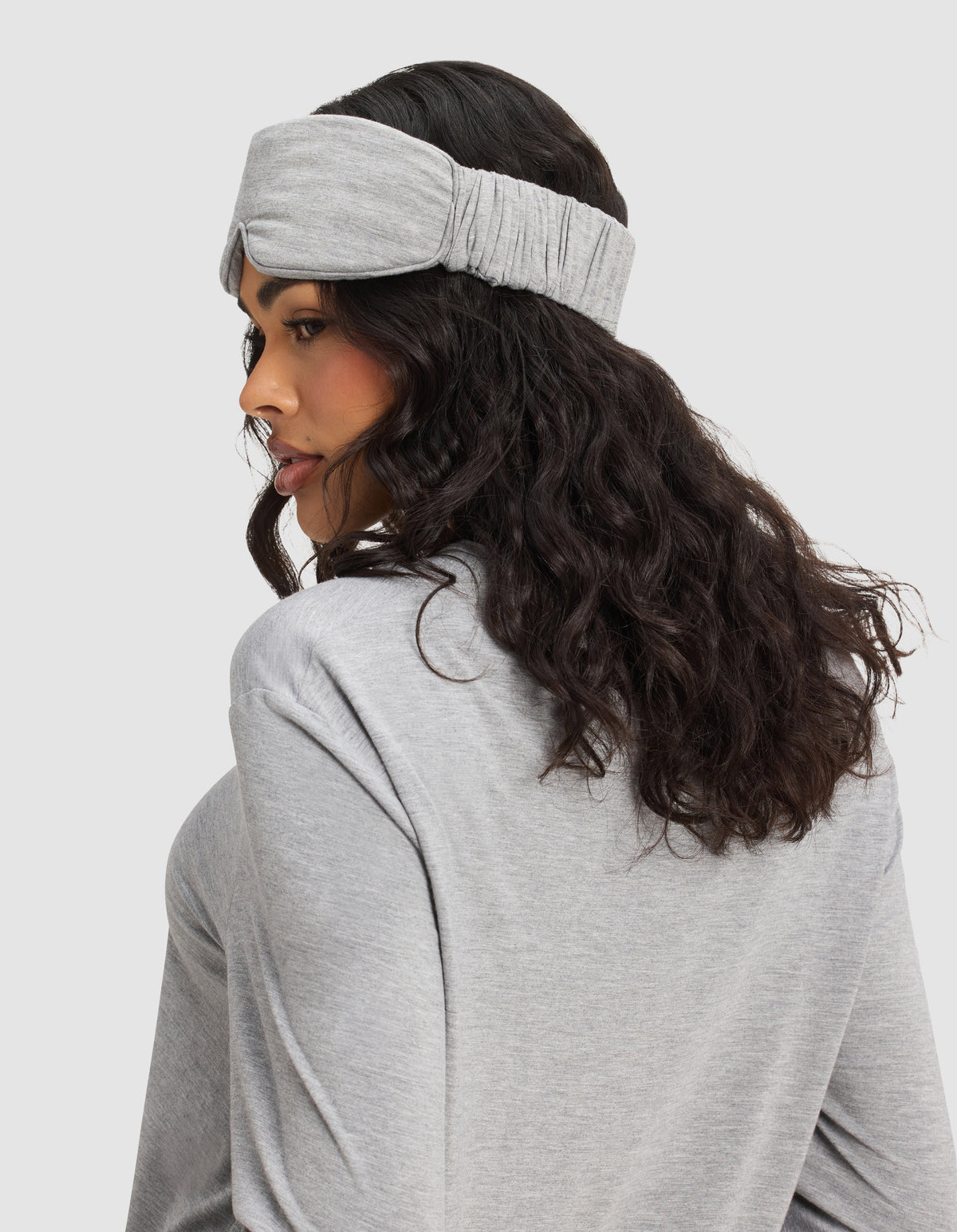 A woman with long, wavy dark hair wears the Cozy Earth Bamboo Stretch-Knit Sleep Mask and a matching gray top, shown in side view against a plain light background.