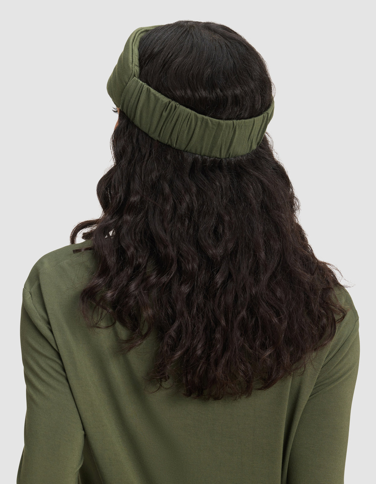 A person with long, wavy dark hair is shown from behind wearing a Cozy Earth Bamboo Stretch-Knit Sleep Mask in olive green, with a matching top against a plain light-colored background.