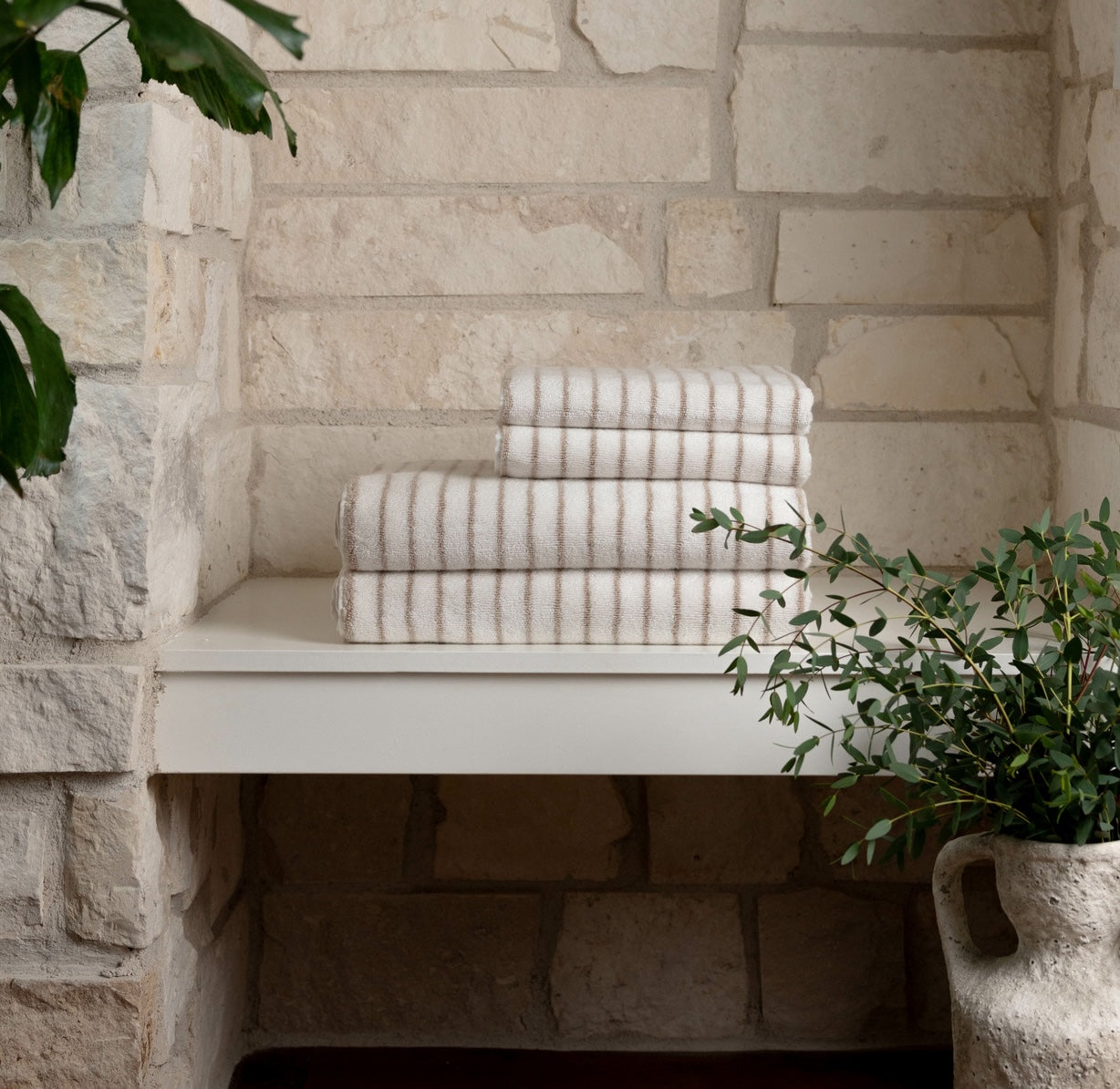 A stack of Cozy Earth Striped Luxe Bath Towels sit on a bench with a stone wall behind and plants nearby