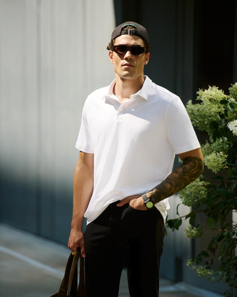 A man wearing a white Cozy Earth Everday Polo shirt, black pants, a black cap, and sunglasses, standing outdoors next to a gray wall and green foliage.