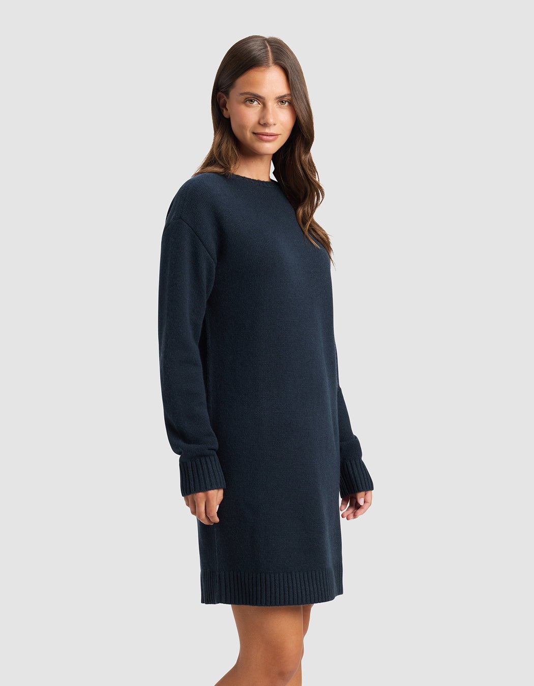 A woman with long brown hair models the Cozy Earth Women's Brooklyn Sweater Dress, a loose-fitting, long-sleeve navy blue piece, standing against a plain light gray background and smiling gently.