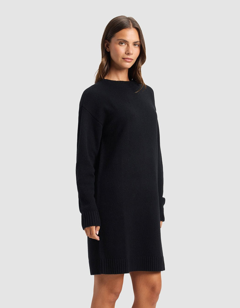 A woman with straight brown hair and a neutral expression stands sideways in a plain light gray setting, modeling the Cozy Earth Women's Brooklyn Sweater Dress—an elegant, long-sleeved, knee-length black dress.