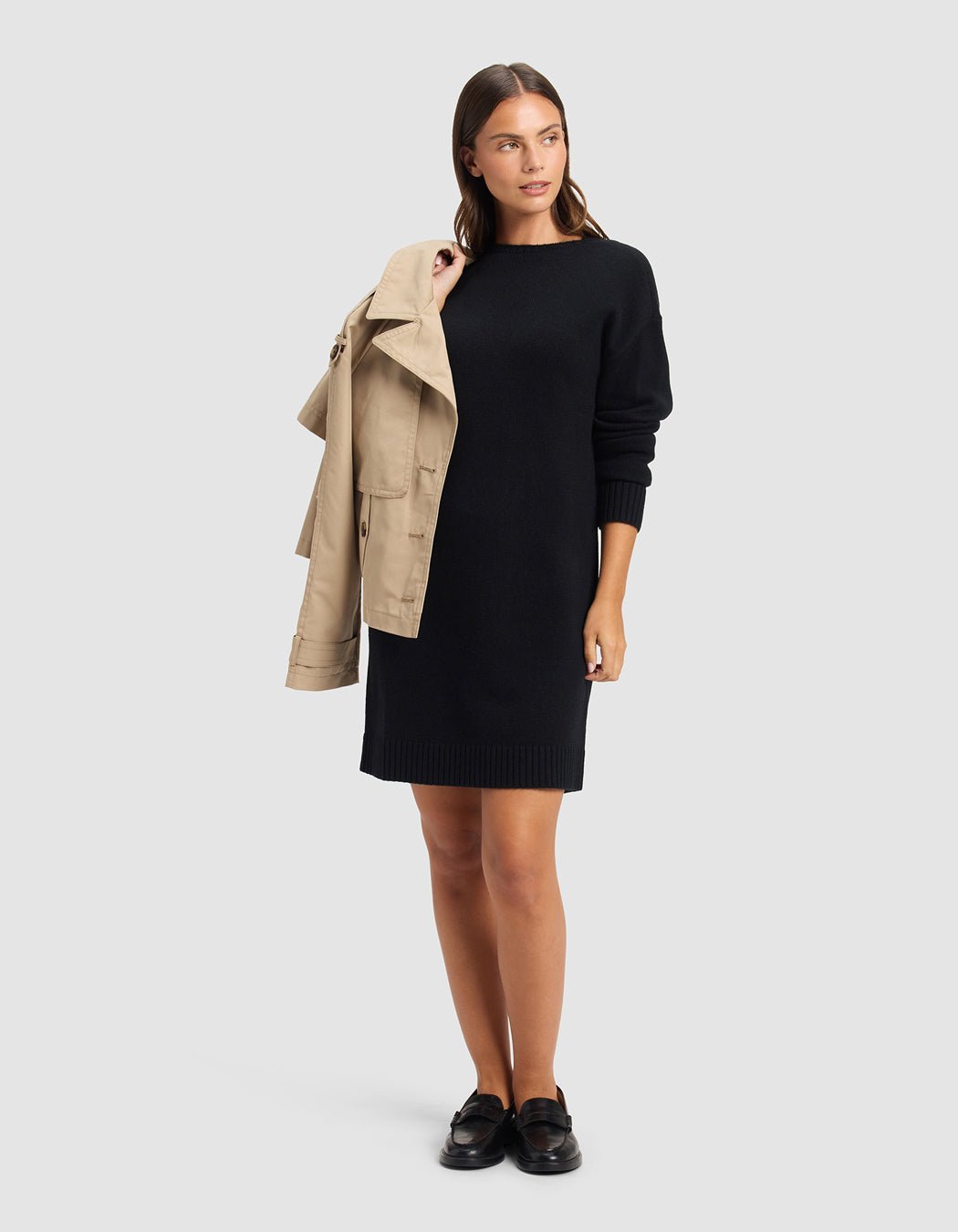 A woman stands against a plain light gray background, wearing Cozy Earth's Women's Brooklyn Sweater Dress in black with black loafers and holding a beige trench coat over her shoulder.