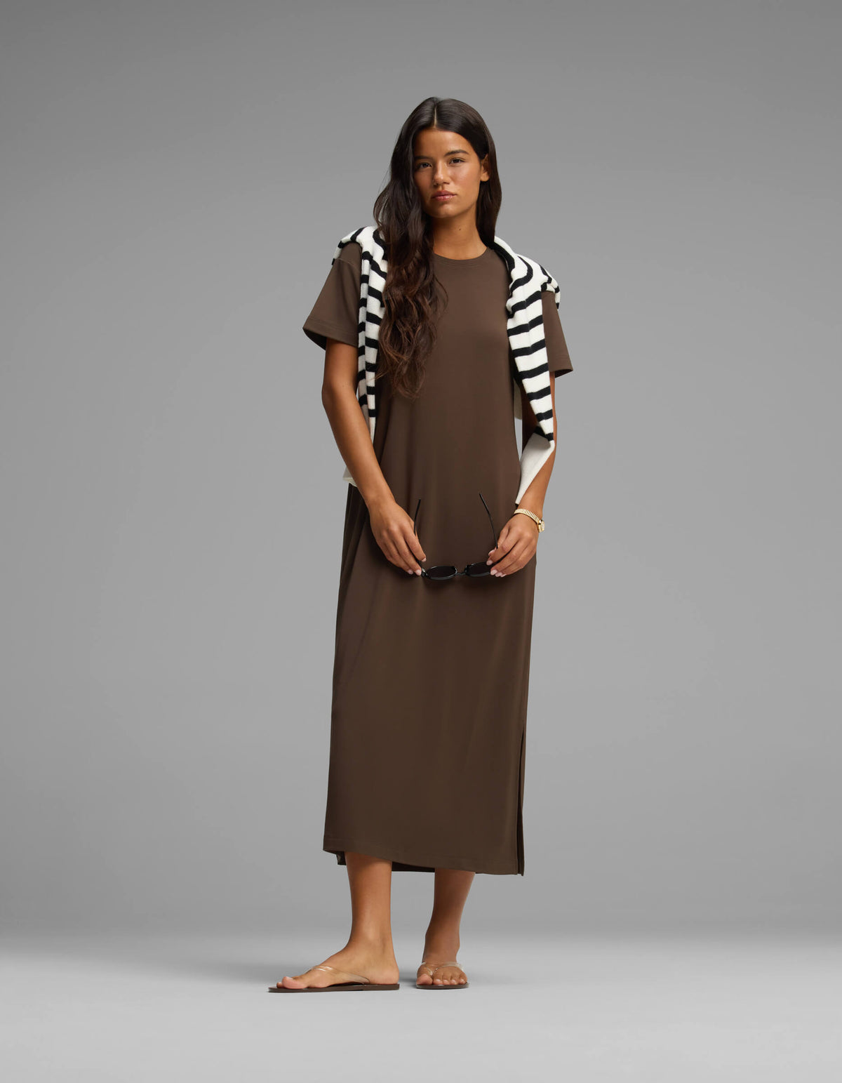 A woman stands barefoot against a plain gray background, wearing the Cozy Earth Women's Brushed Bamboo Midi Dress in brown with a black and white striped sweater over her shoulders, holding sunglasses in one hand.