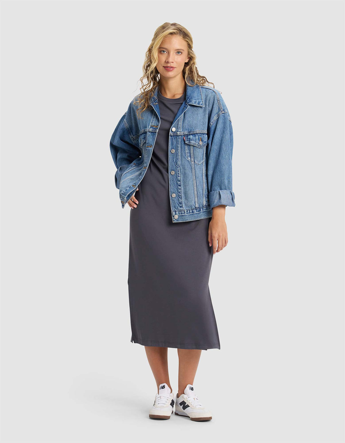 A woman with wavy blonde hair and a confident expression wears the Cozy Earth Women's Brushed Bamboo Midi Dress in dark gray, styled with a blue denim jacket and white sneakers against a plain background.