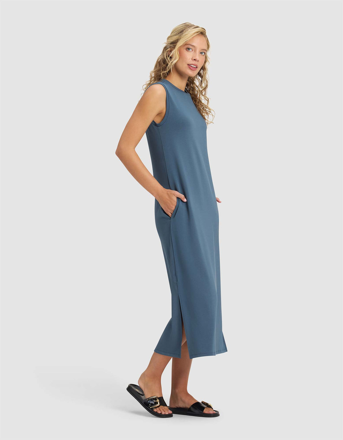 A woman with blonde hair wears the Cozy Earth Women's Brushed Bamboo Sleeveless Midi Dress in blue with side pockets and black slide sandals, standing sideways and smiling against a plain white background.