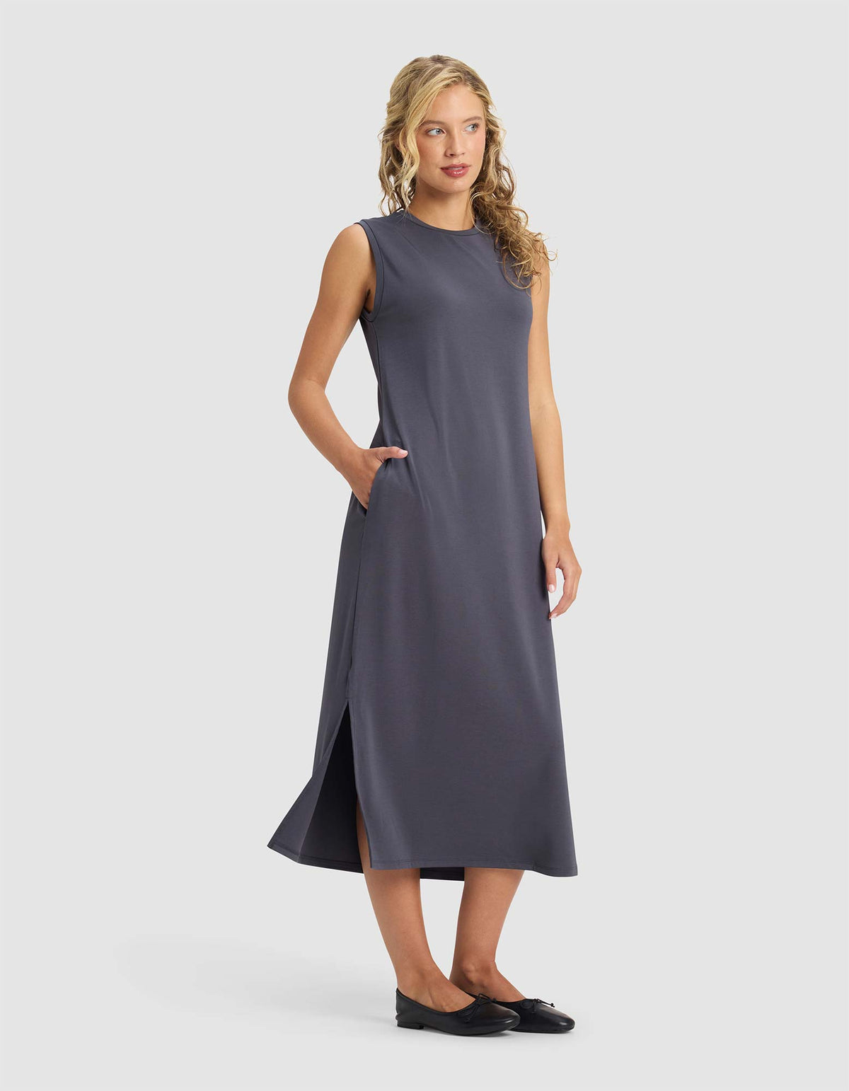 A woman with wavy blonde hair models the Cozy Earth Women's Brushed Bamboo Sleeveless Midi Dress in dark gray, featuring side pockets and a side slit, styled with black flats against a plain light gray background.