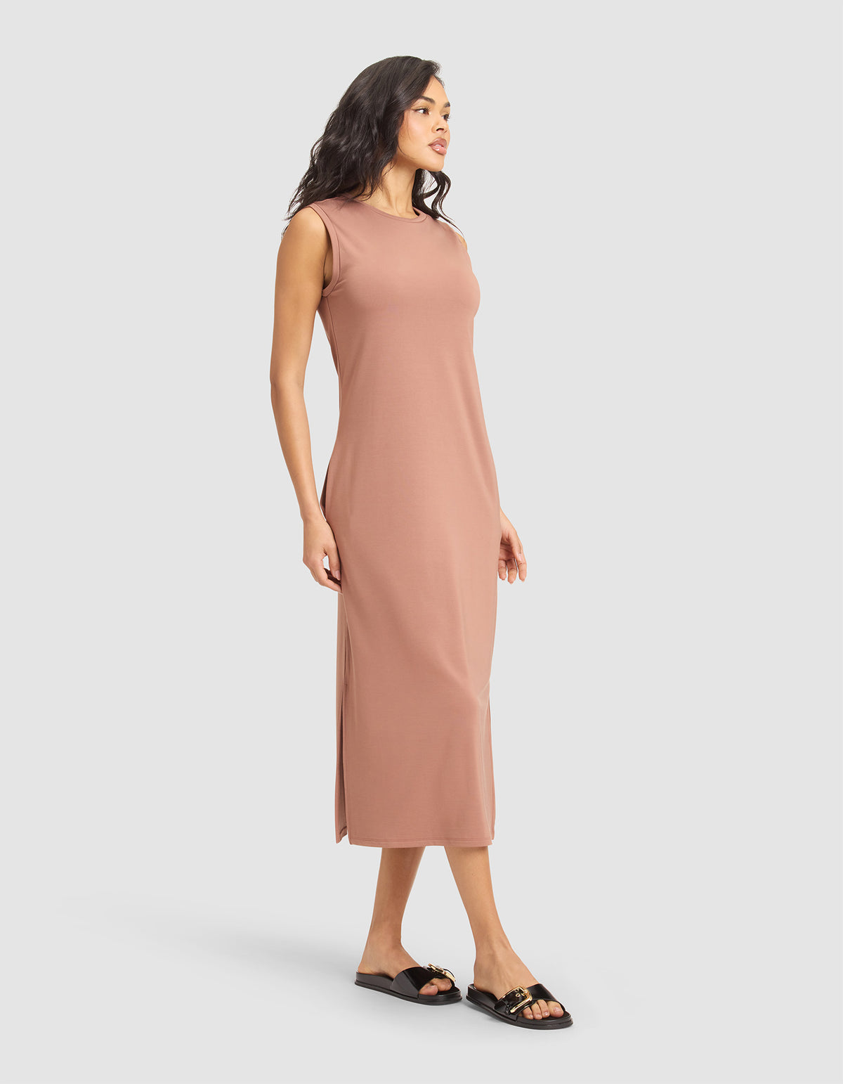 A woman with medium-length dark hair wears the Cozy Earth Women's Brushed Bamboo Sleeveless Midi Dress in light brown, featuring side slits and a midi length. She pairs it with black slide sandals against a plain white background. |Color:Saddle
