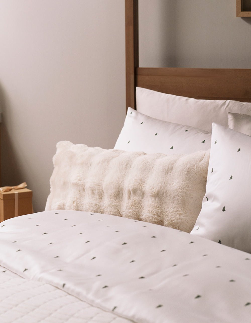 A neatly made bed with white sheets and tree-patterned pillowcases, accented by the Cozy Earth Bubble Cuddle Pillow in cream, set against a wooden headboard in a softly lit room.