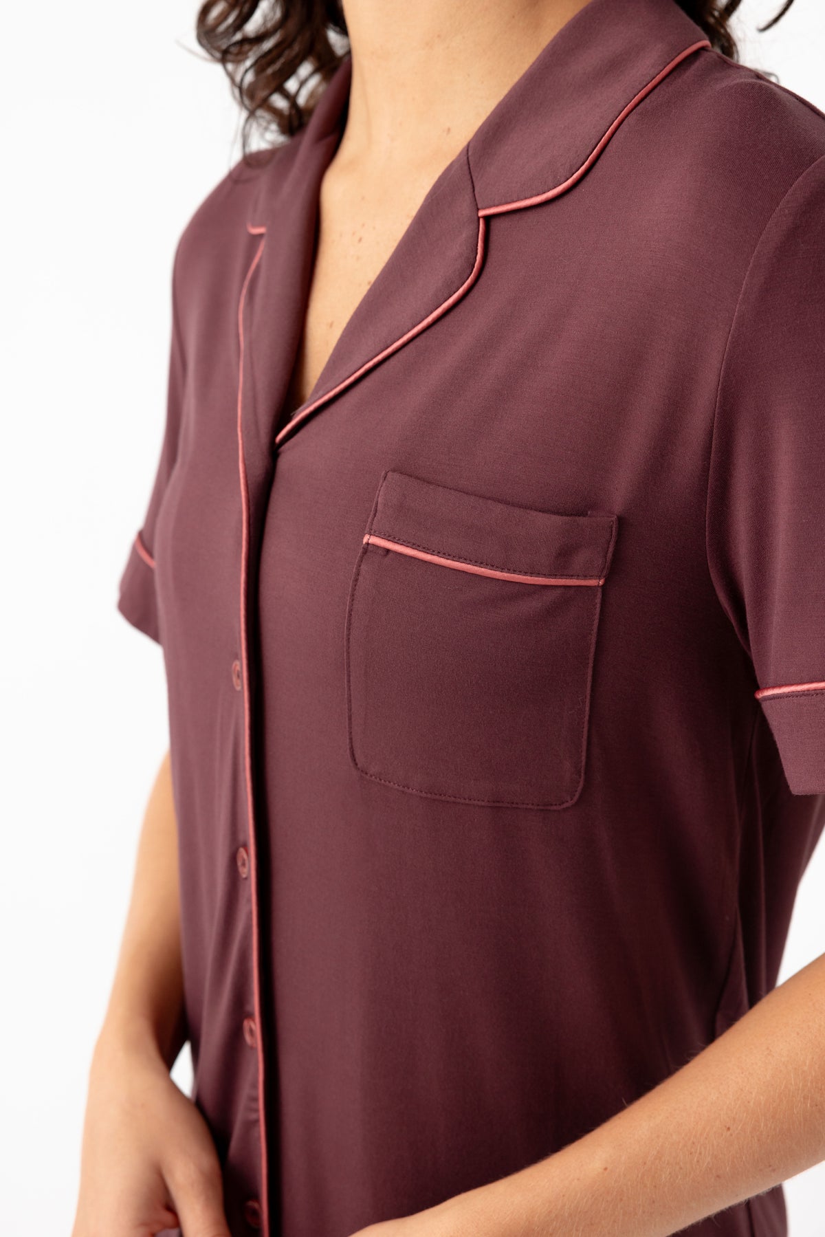 A person wears the Cozy Earth Women's Bamboo Stretch-Knit Short Sleeve Pajama Set—a maroon, pink-piped, button-down top with a breast pocket. The person's head is partially out of frame.
