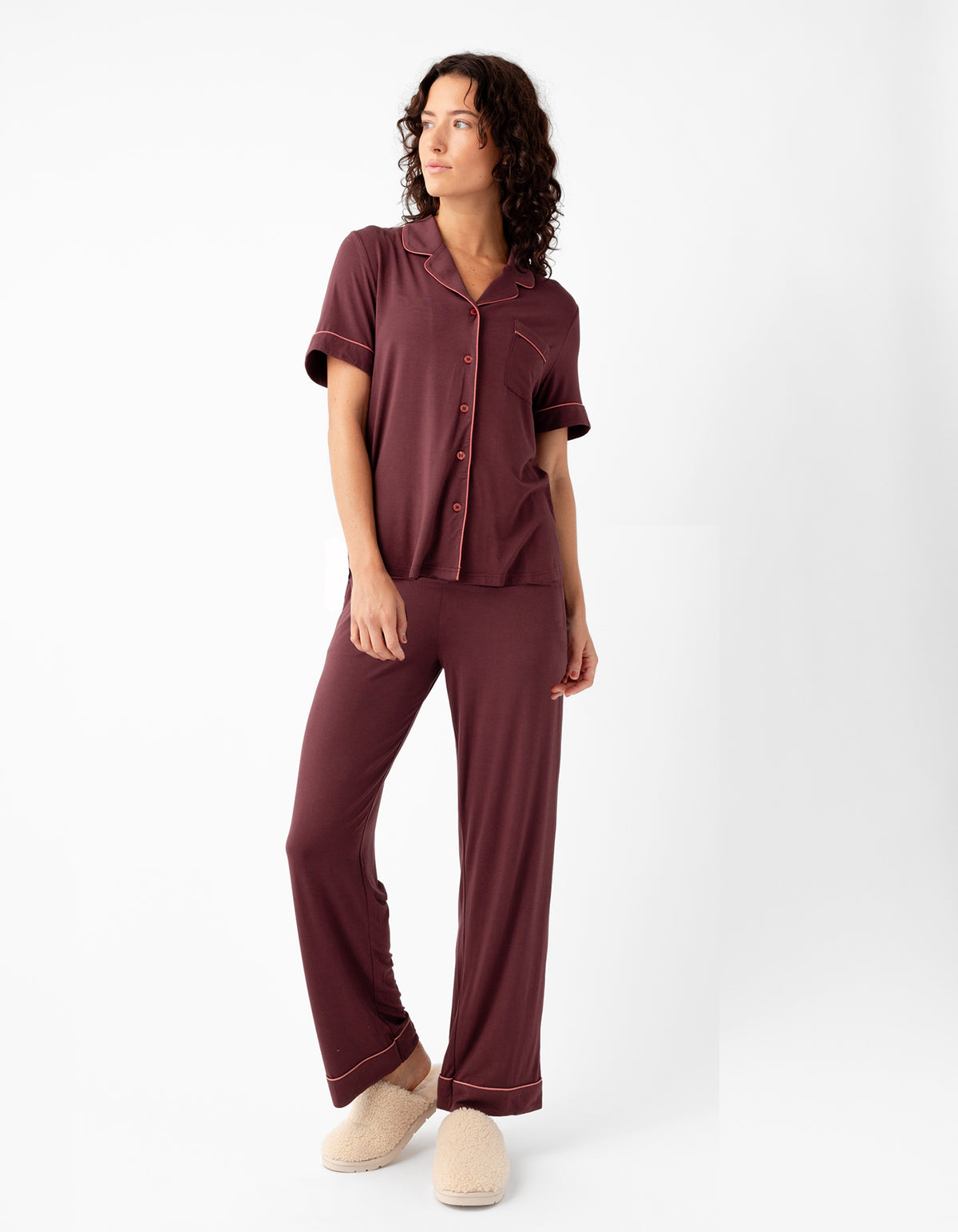A woman in Cozy Earth's Women's Bamboo Stretch-Knit Short Sleeve & Pant Pajama Set, featuring a collar, buttons, and loose-fitting pants in burgundy, stands against a white background wearing beige slippers and looking to the side with one hand on her hip.