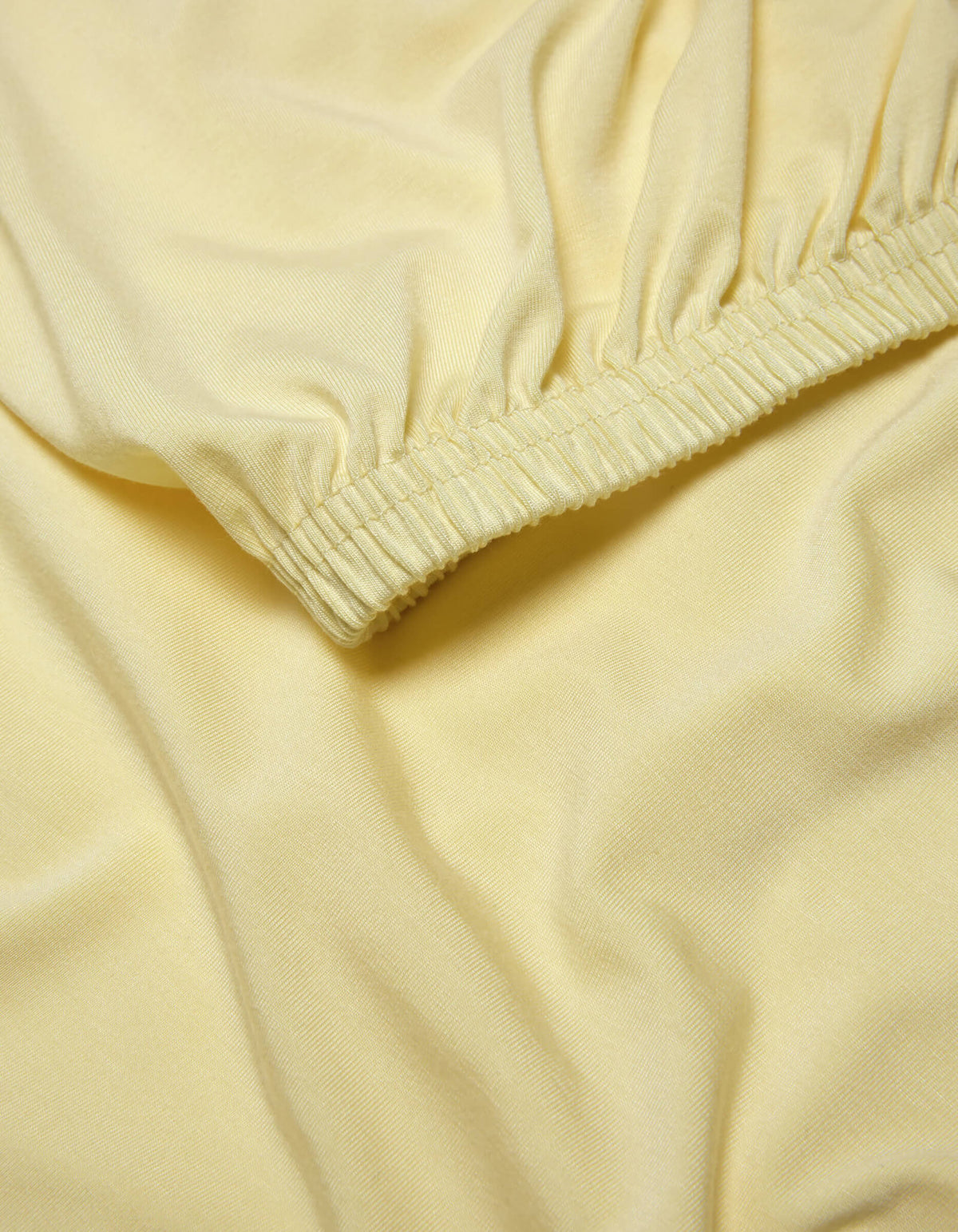 A close-up of the Cozy Earth Bamboo Jersey Fitted Sheet shows light yellow fabric with elasticized, gathered edges that create gentle folds and a smooth, soft texture.
