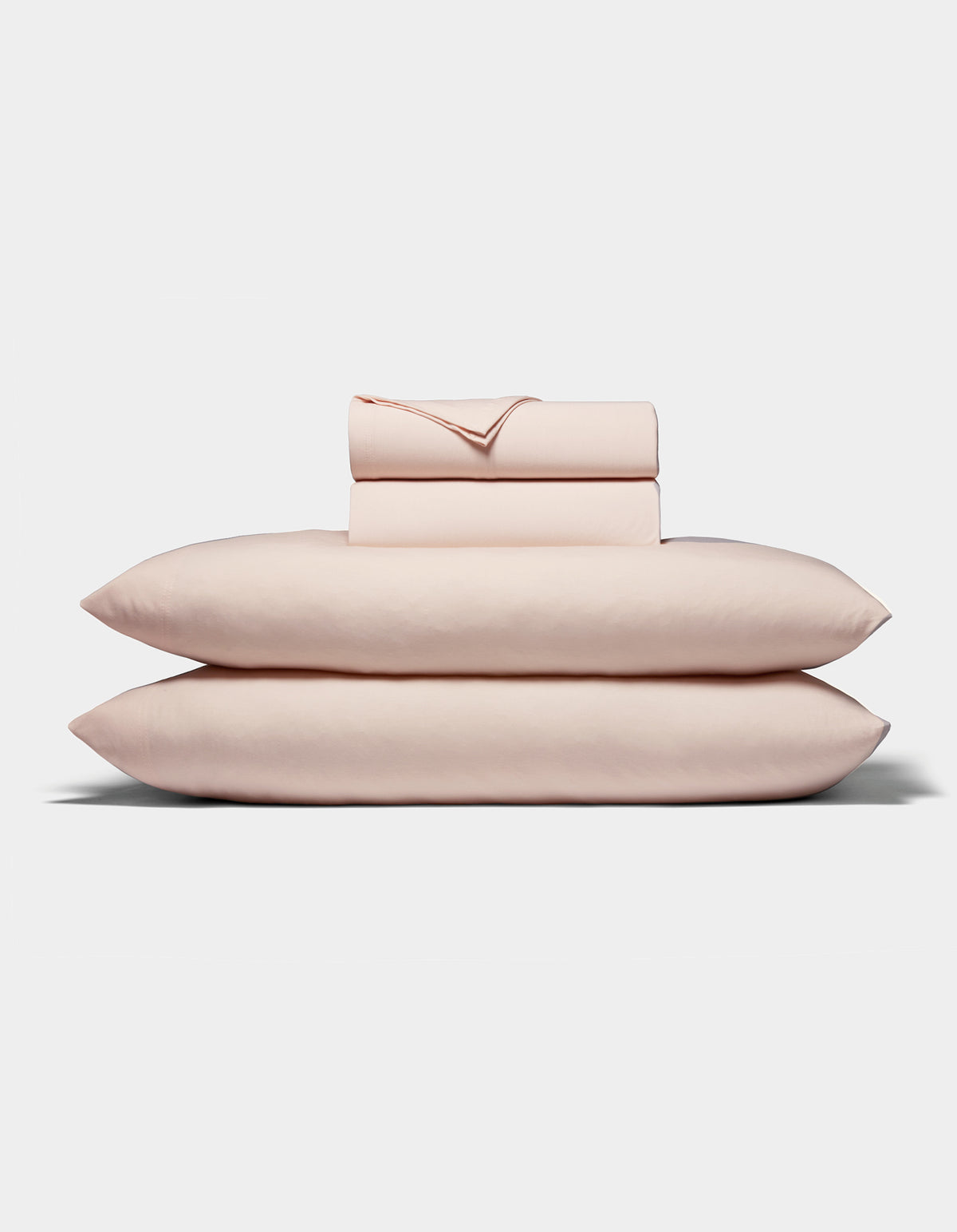 A neatly folded stack of Cozy Earth's Bamboo Jersey Sheet Set, in pale pink, featuring two pillows, a fitted sheet, a flat sheet, and pillowcases arranged on a white background.