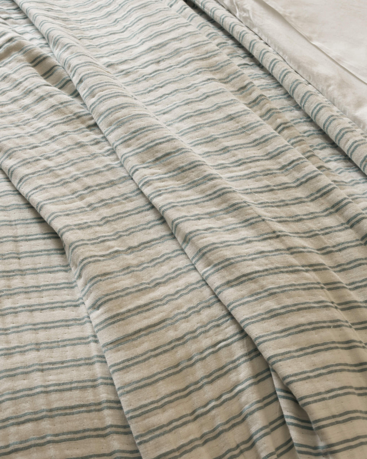 The Waterfront Coverlet by Cozy Earth is shown on a bed, displaying its cream color with thin blue-green stripes and gentle folds. A smooth white sheet is visible at the top right corner.