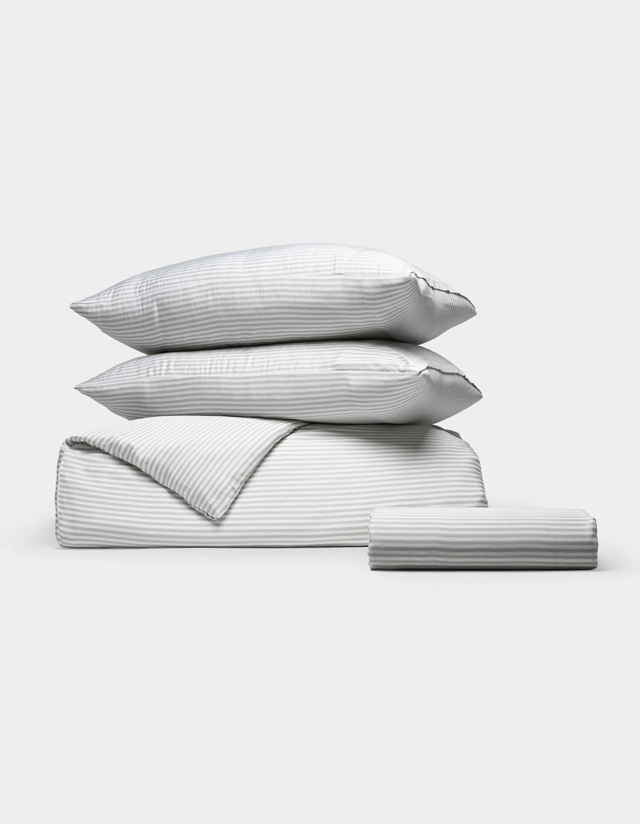 The Cozy Earth Bamboo Bedding Essential Bundle, featuring two stacked pillows, a folded comforter, and a folded sheet in light gray and white thin stripes, is arranged neatly against a plain white background.|Color:Classic Stripe in Dew