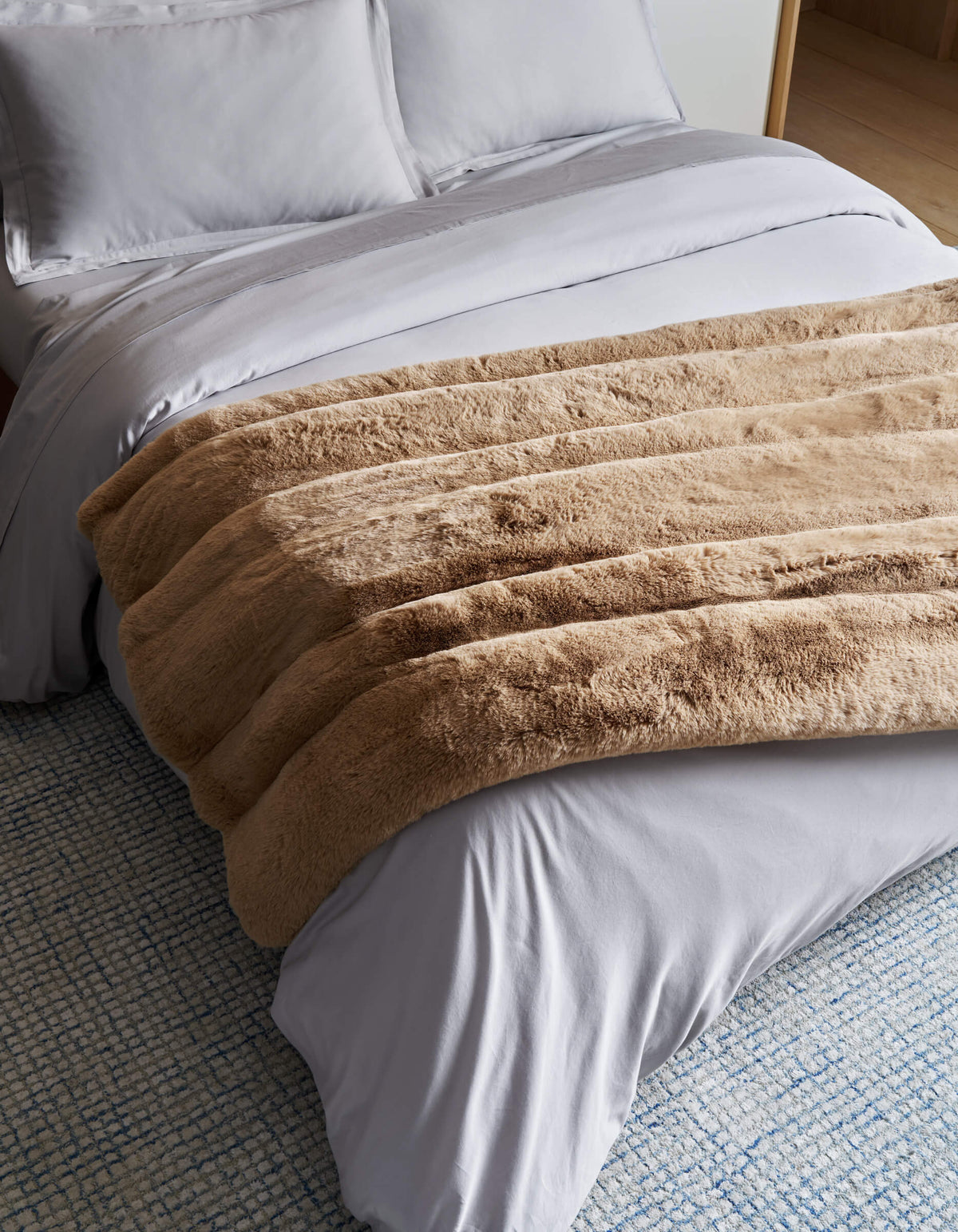 A neatly made bed with white sheets and pillows is topped with the Cozy Earth Channel Cuddle Blanket in thick tan faux fur, set on a textured blue and white rug.