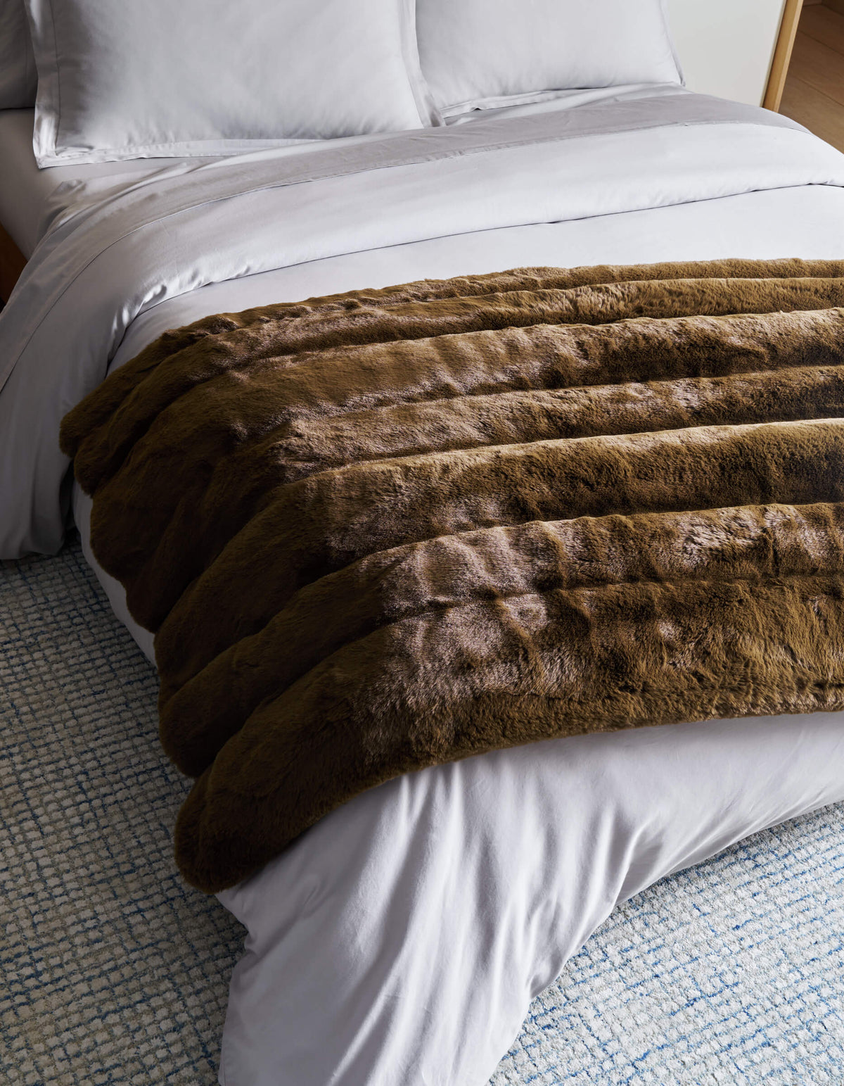 A neatly made bed with white sheets and pillows features the plush Cozy Earth Channel Cuddle Blanket in brown draped across the foot, on top of a blue and white textured rug.