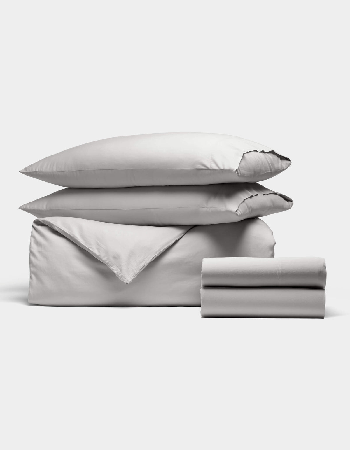The Soft-Wash Cotton Bedding Deluxe Bundle by Cozy Earth features a neatly stacked set of light gray bedding—two pillows, a folded duvet, and two folded sheets—arranged against a plain white background.