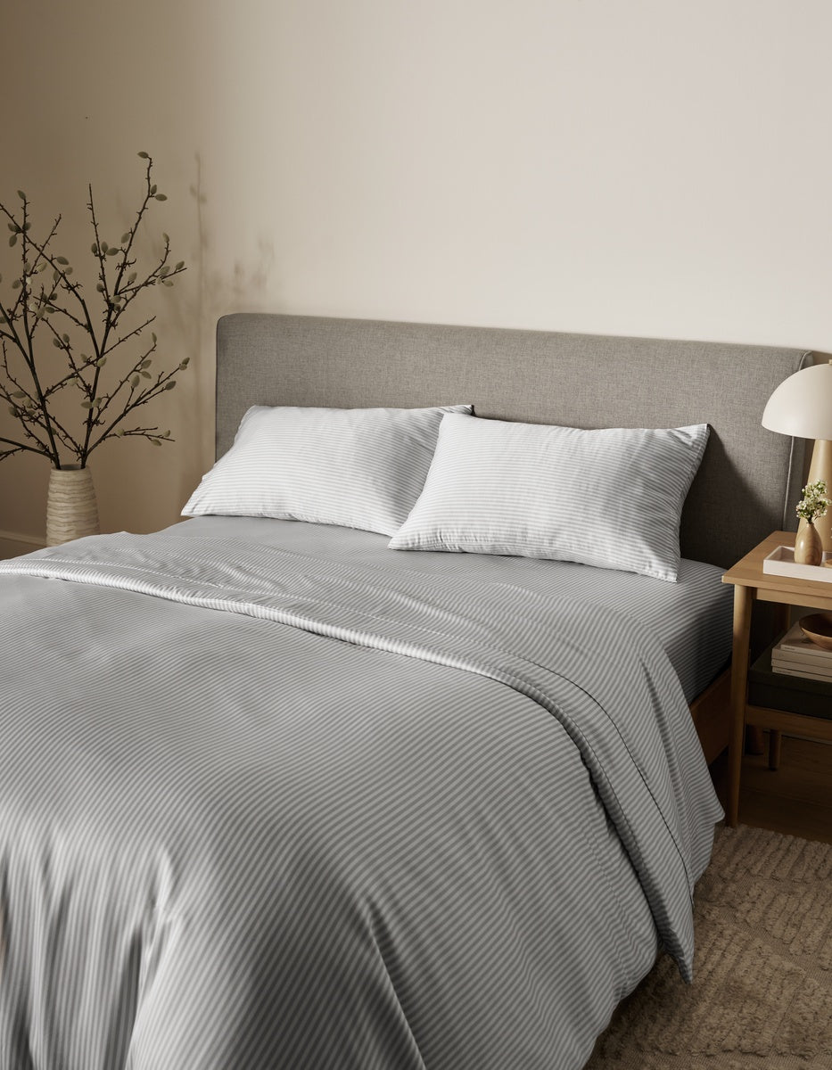 A neatly made bed in a minimalist bedroom setting, featuring a gray and white vertically striped duvet cover and two white pillows against a textured gray headboard.|Color:Classic Stripe in Dew