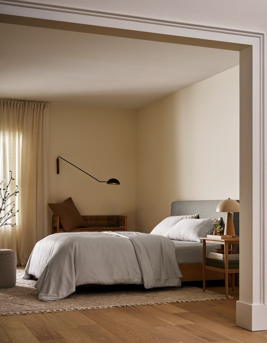 A minimalist bedroom interior with a neatly made bed dressed in white linens, a gray upholstered headboard, warm beige walls, wood flooring, a bedside table with a lamp, and a wall-mounted black lamp. |Color:Classic Stripe in Dew