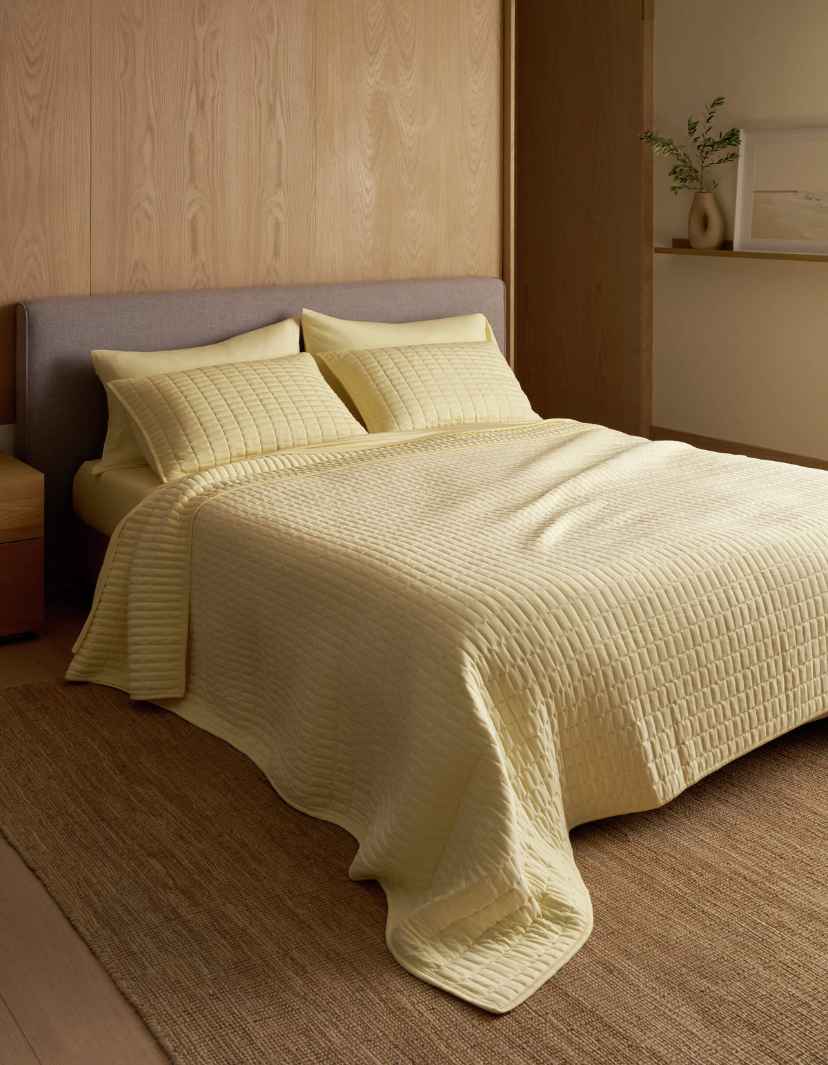 A minimalist bedroom features a neatly made bed with the Cozy Earth Bamboo Jersey Quilt in pale yellow, matching pillows, wood-paneled walls, a neutral rug, and a small shelf displaying a vase and framed art.