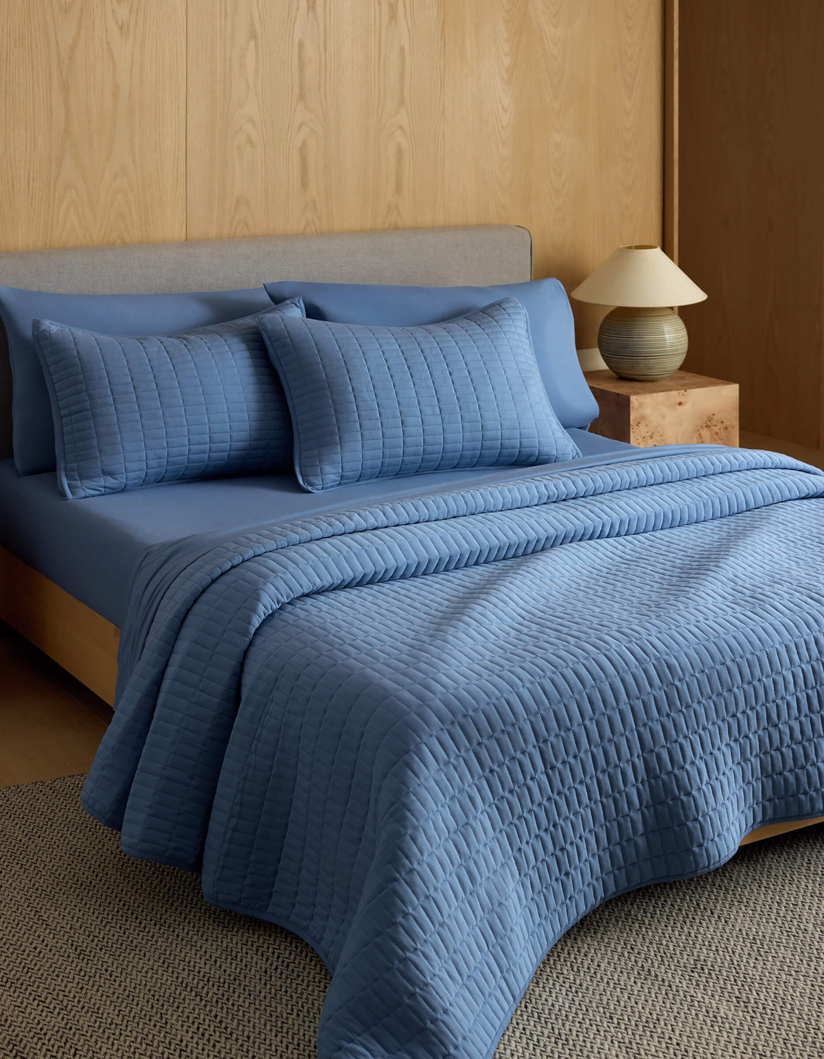 A neatly made bed features the Cozy Earth Bamboo Jersey Quilt with matching pillows, set against a light wood-paneled wall. A small nightstand with a lamp sits beside the bed on a textured rug.|Color:Sailor