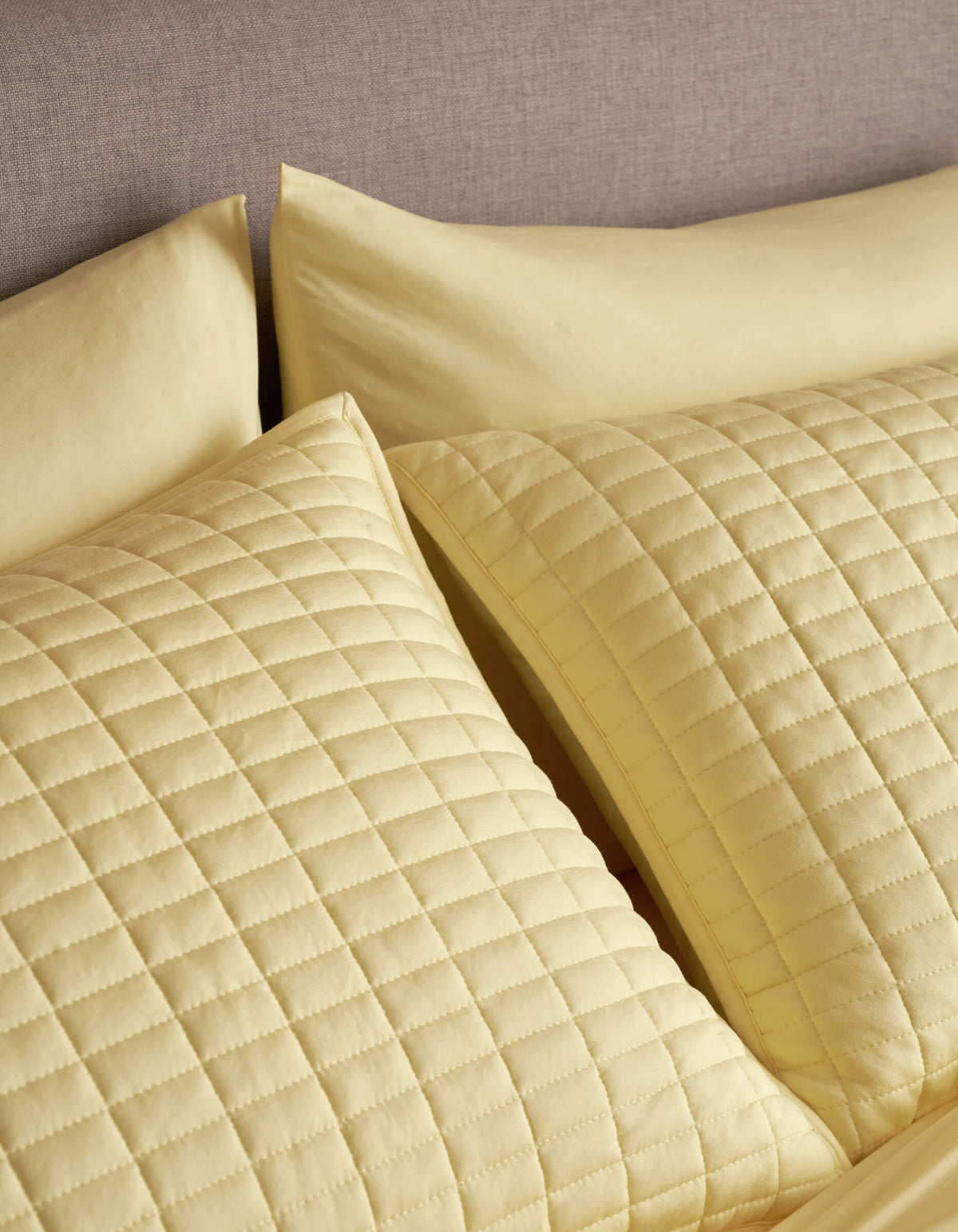 Two Cozy Earth Bamboo Jersey Quilted Shams in yellow, featuring a square pattern, rest against a grey upholstered headboard, paired with matching yellow bedsheets and pillowcases.
