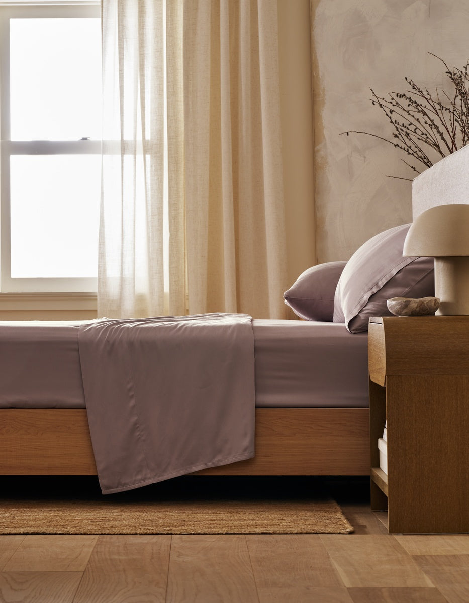 A neatly made bed is dressed in Cozy Earth’s Bamboo Flat Sheet, paired with soft pillows and positioned next to a wooden nightstand topped with a lamp and decorative branches. Light filters through sheer curtains, creating a calm, cozy bedroom atmosphere.|Color:Lavender