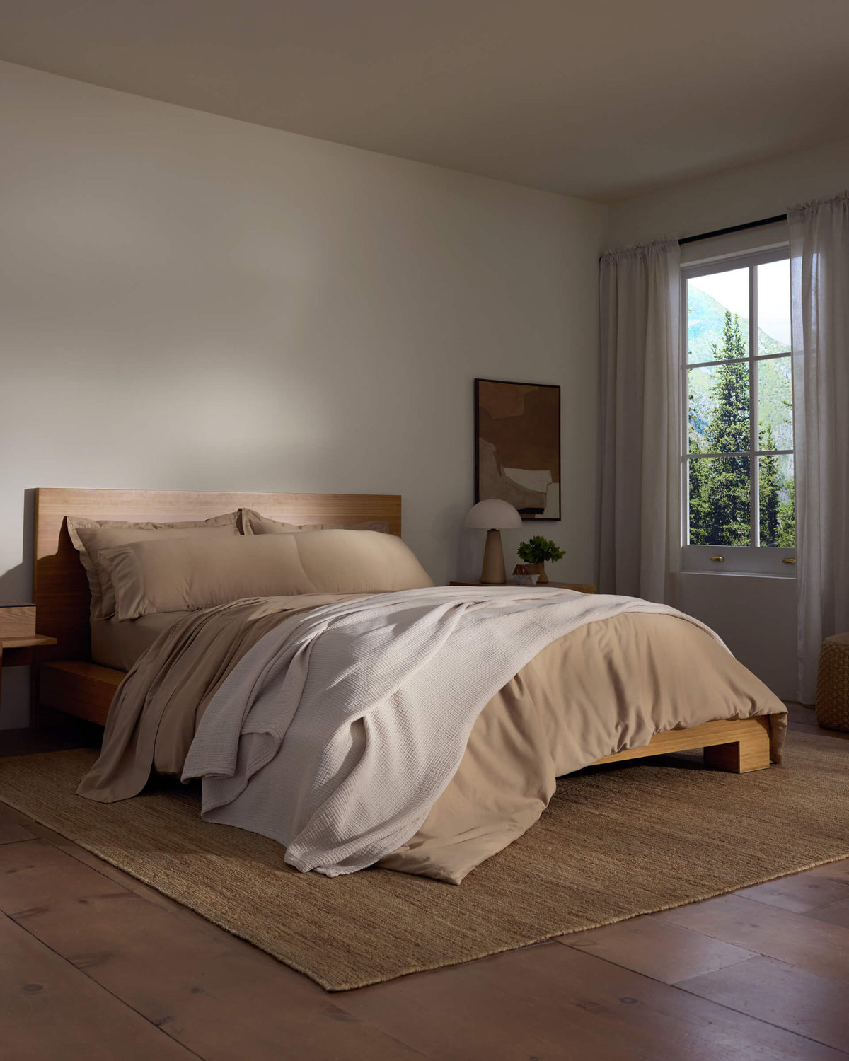 A cozy bedroom features a wooden bed with beige bedding and a white Cotton Gauze Blanket by Cozy Earth, a bedside table with a lamp, sheer curtains letting in natural light, and a woven rug on the wooden floor.